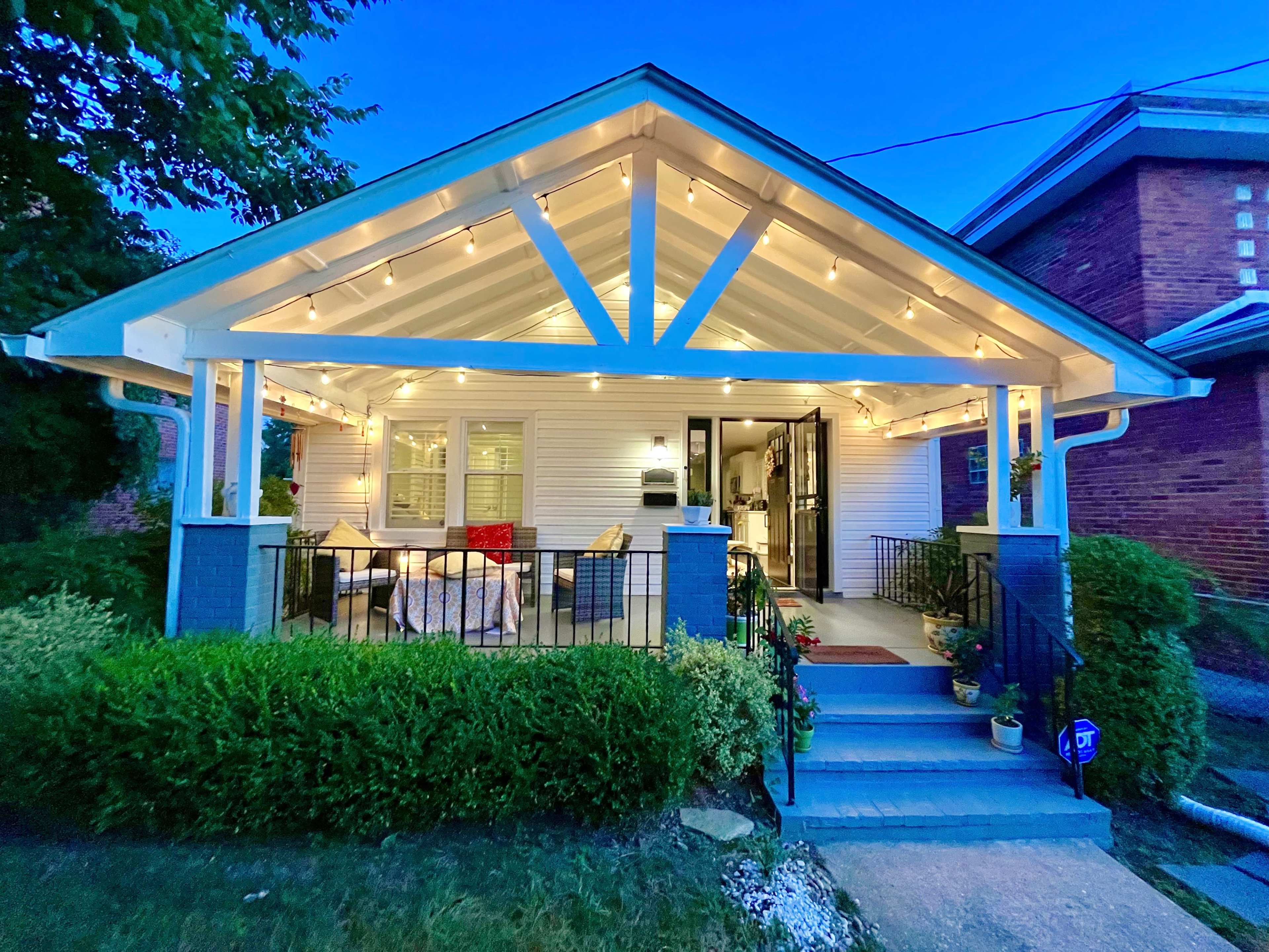 A well-lit porch of a house with decorative lights, surrounded by greenery and a welcoming entrance.