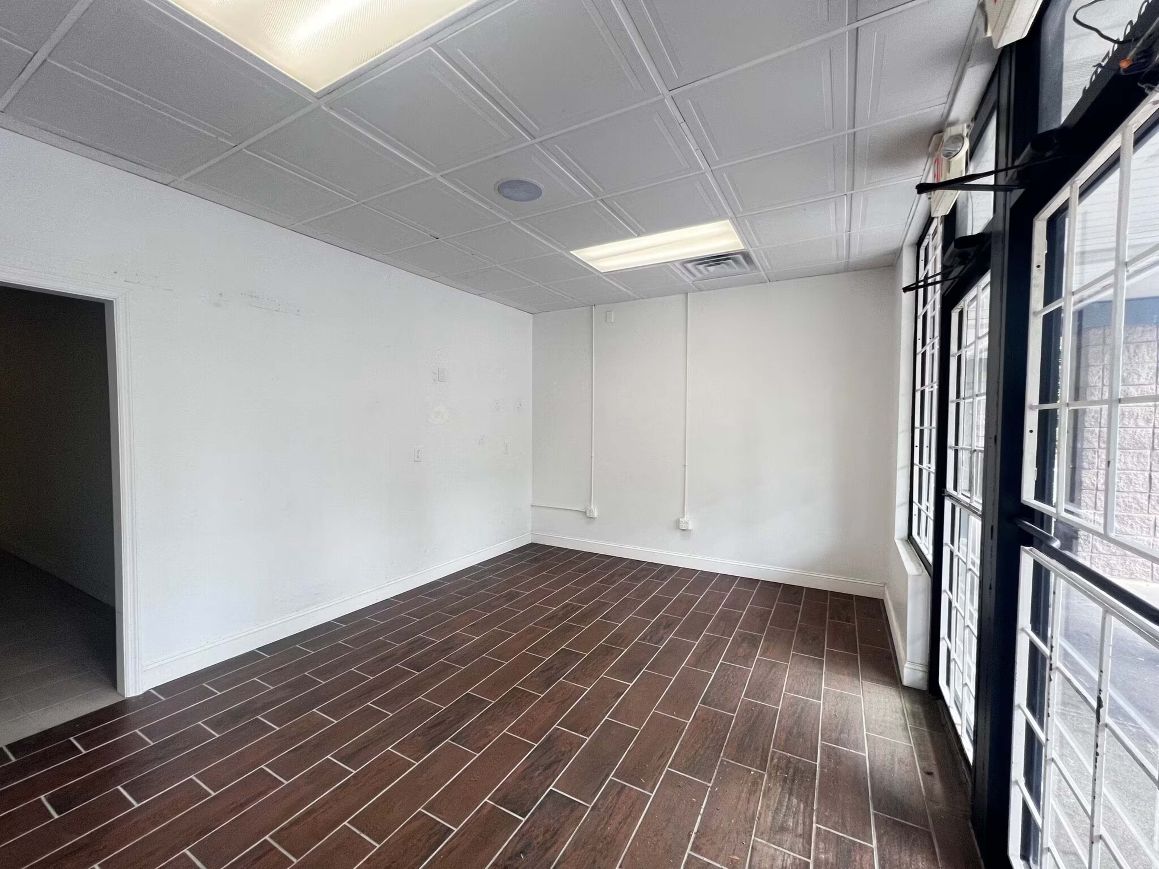 The image shows an empty room with brown tiled flooring, white walls, and large windows on one side.