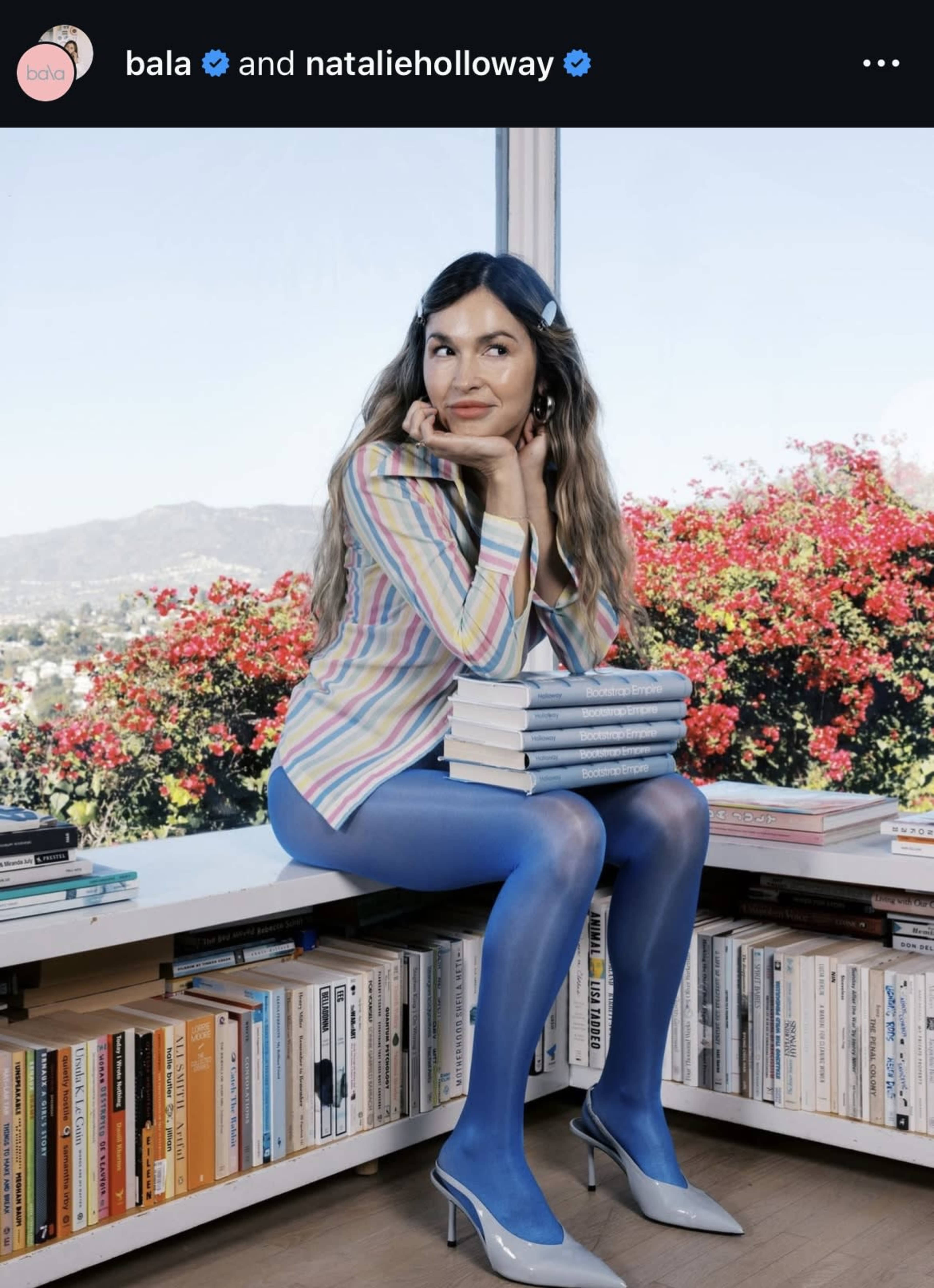 A woman in a striped shirt and blue tights sits on a white shelf with stacks of books, surrounded by a backdrop of blooming flowers and a city skyline.