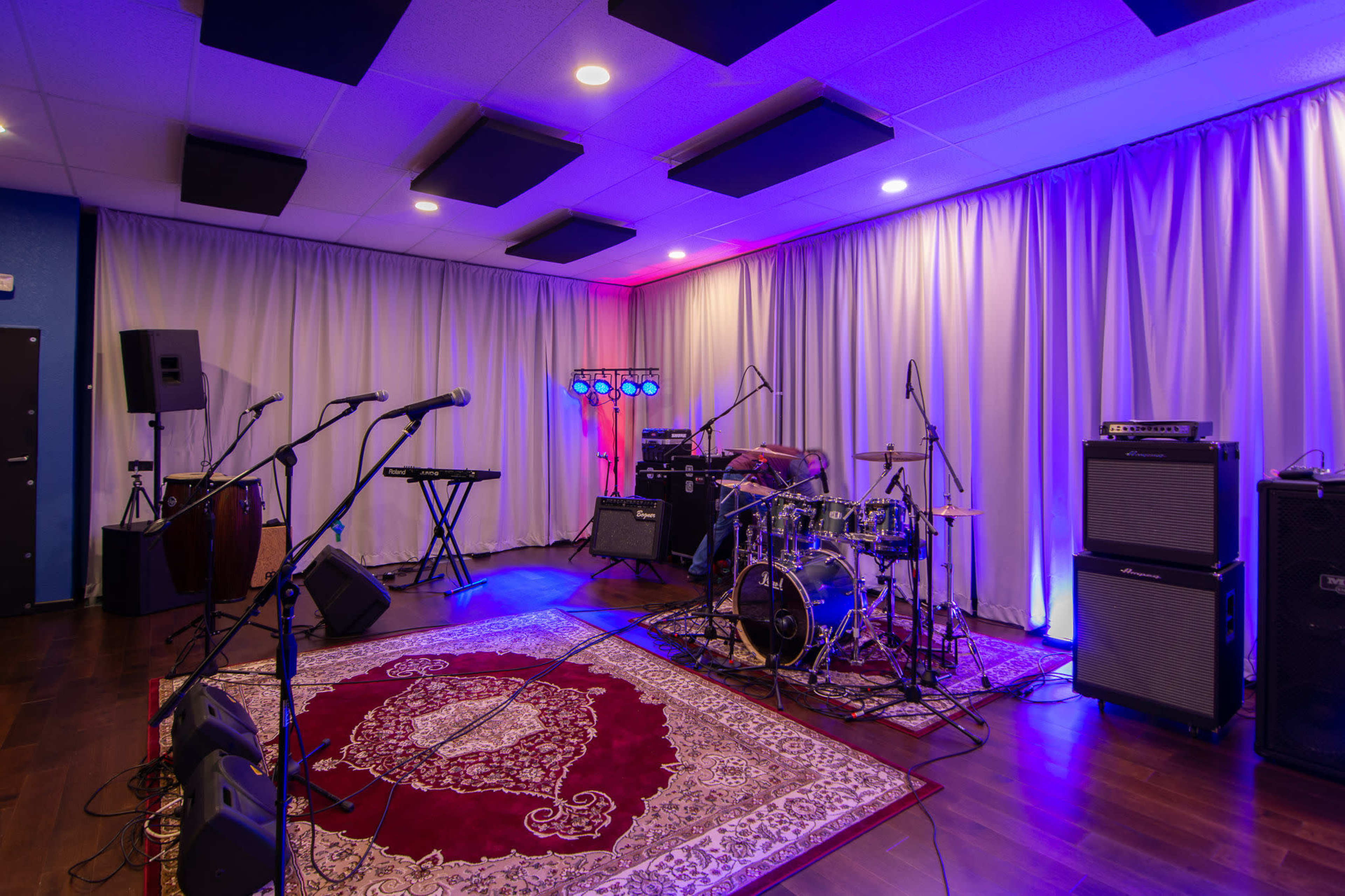 A music rehearsal space featuring a drum set, amplifiers, microphones, and a keyboard, all set against a backdrop of colorful lighting and curtains.