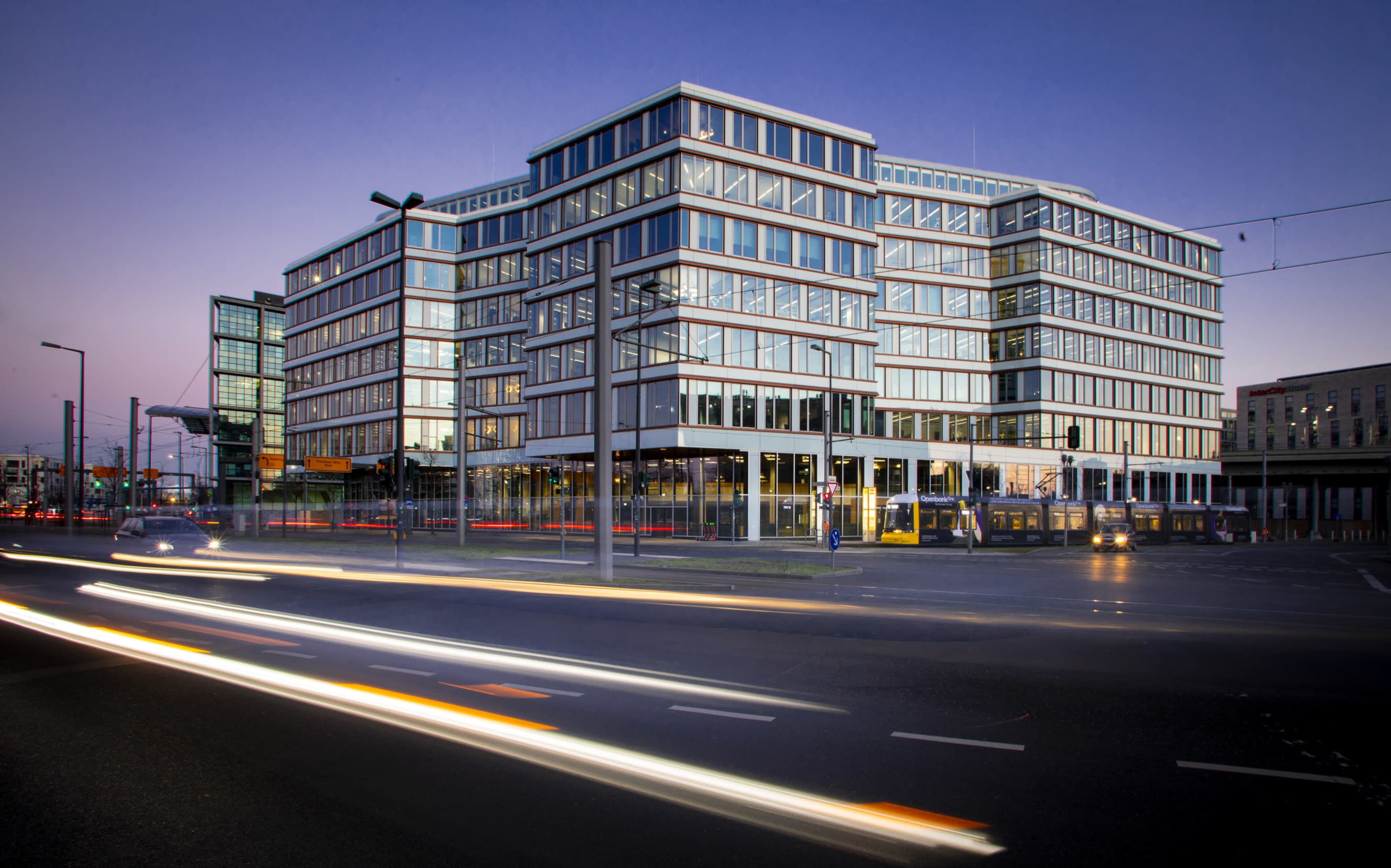A modern office building with large glass windows is situated at a busy intersection, illuminated by dusk light and passing vehicle headlights.