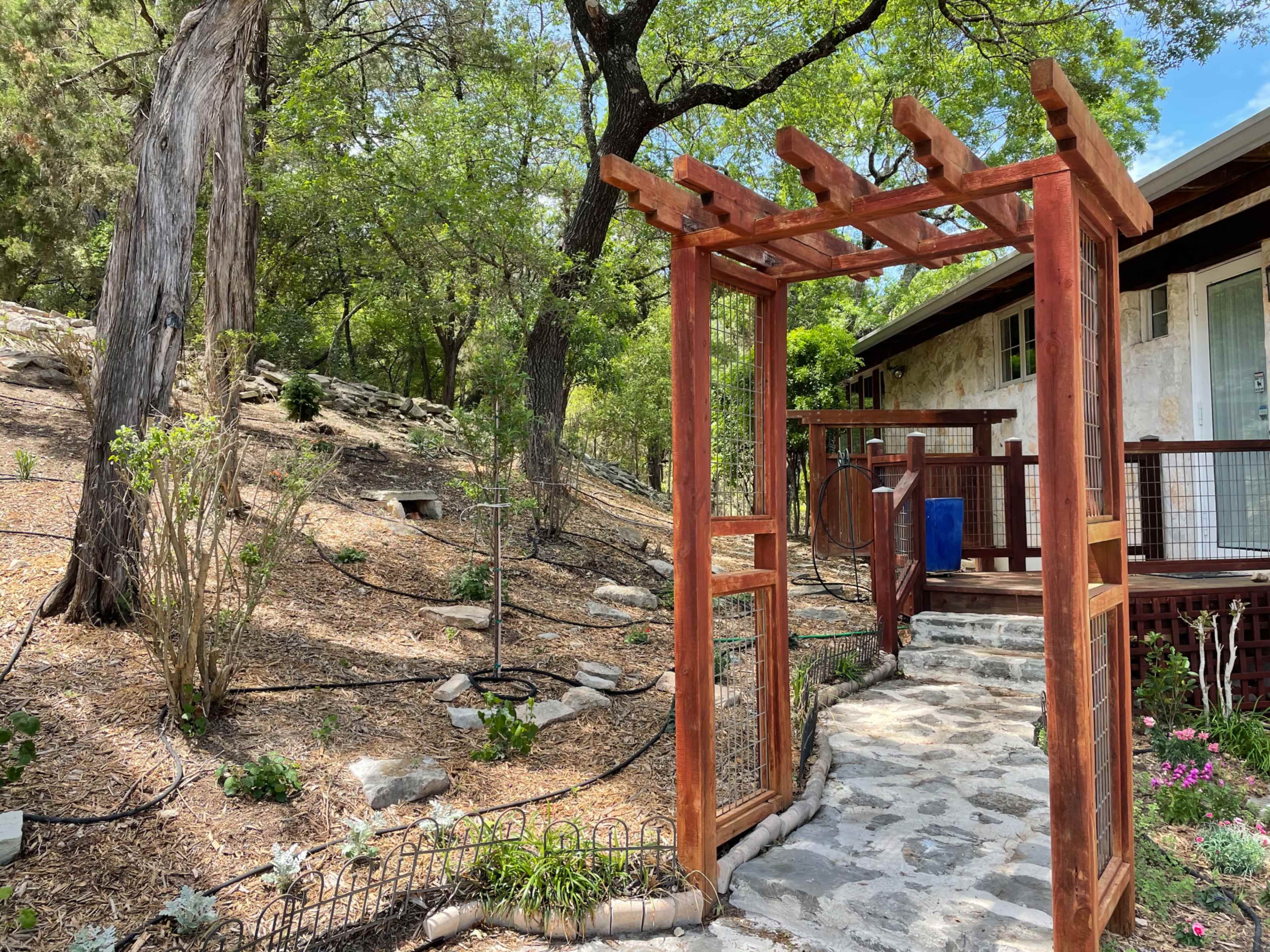 A wooden trellis frames the entrance to a stone pathway leading to a house amidst a landscaped garden with trees and shrubs.