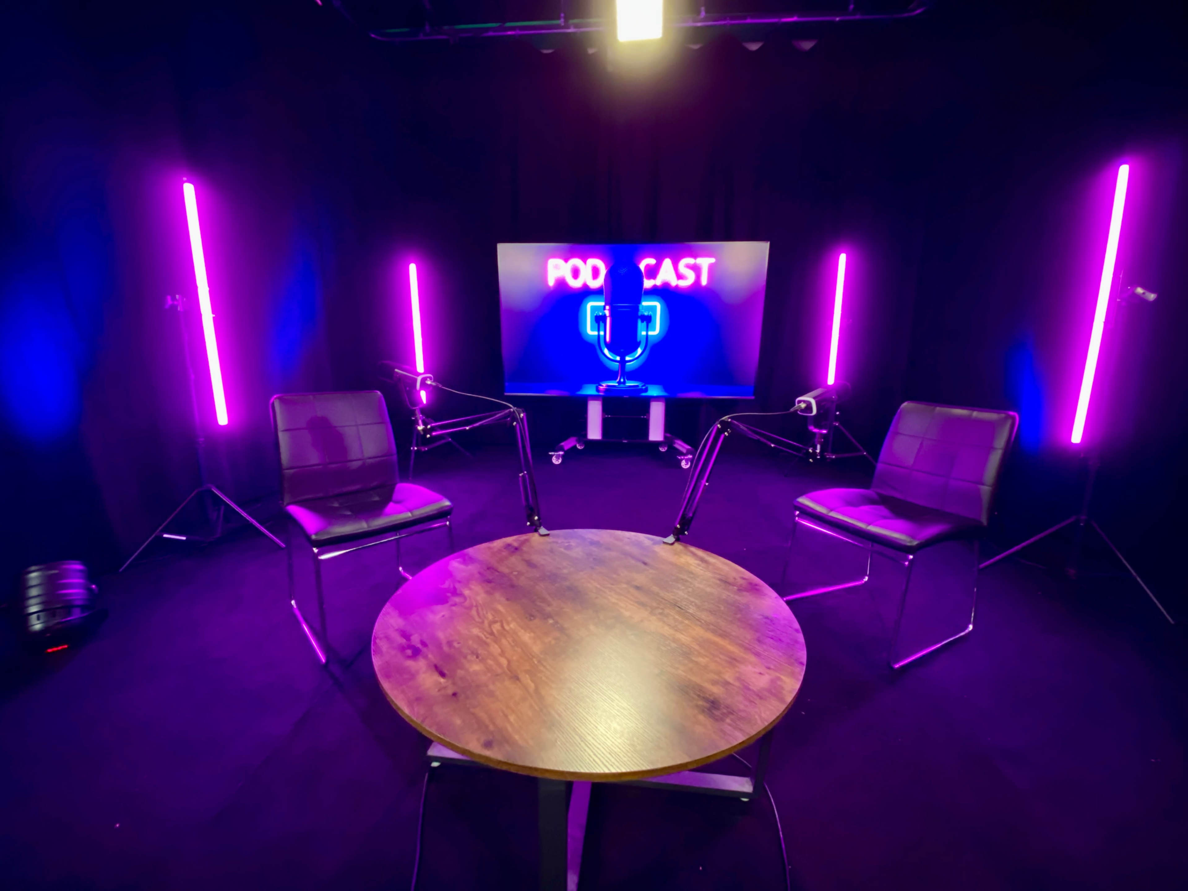 The image shows a podcast studio setup with two chairs facing each other, a round table, and a screen displaying the word "PODCAST" illuminated by pink and blue lights.