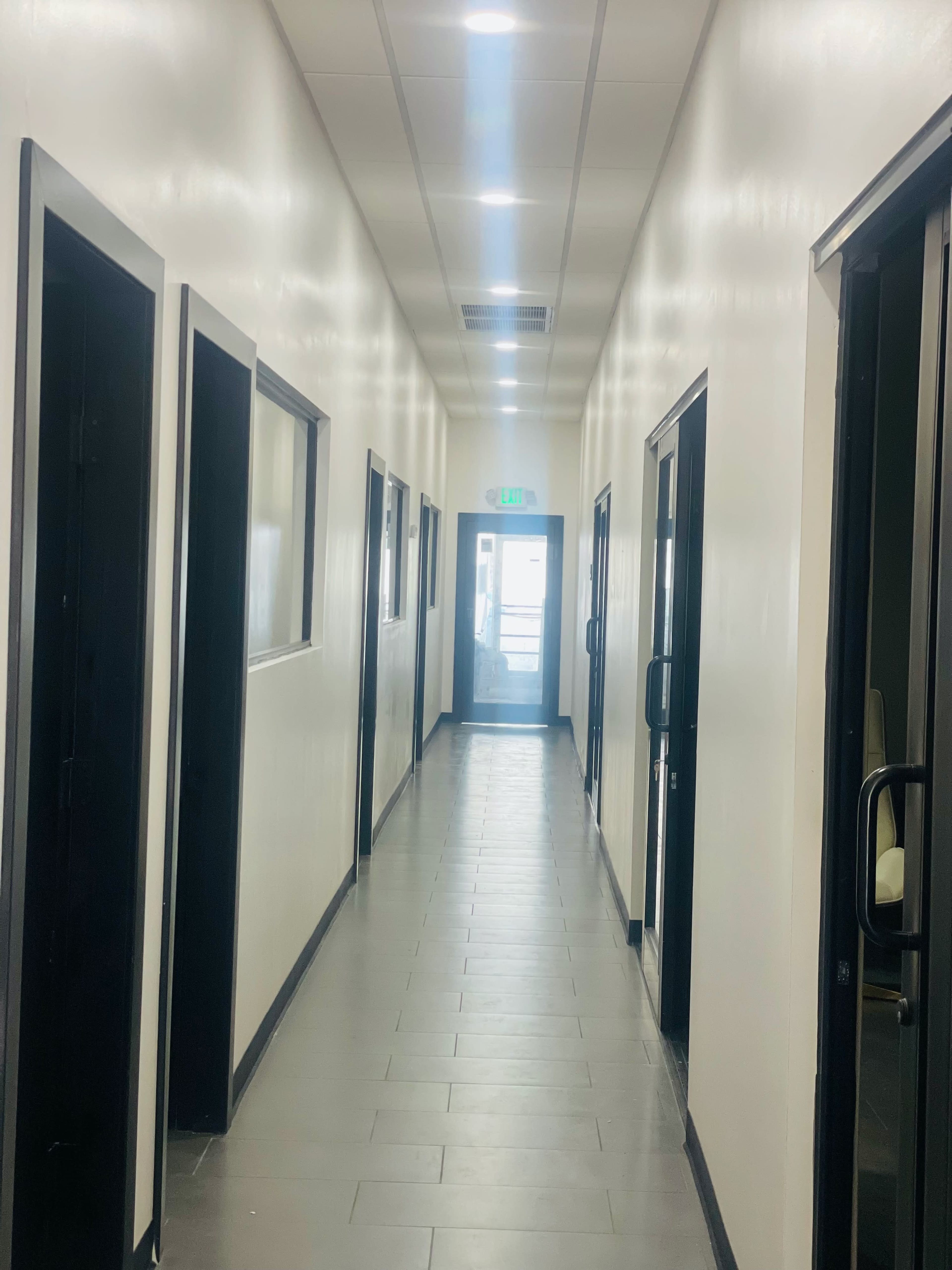 The image shows a long, narrow hallway with several doors on each side and a glass door at the end.