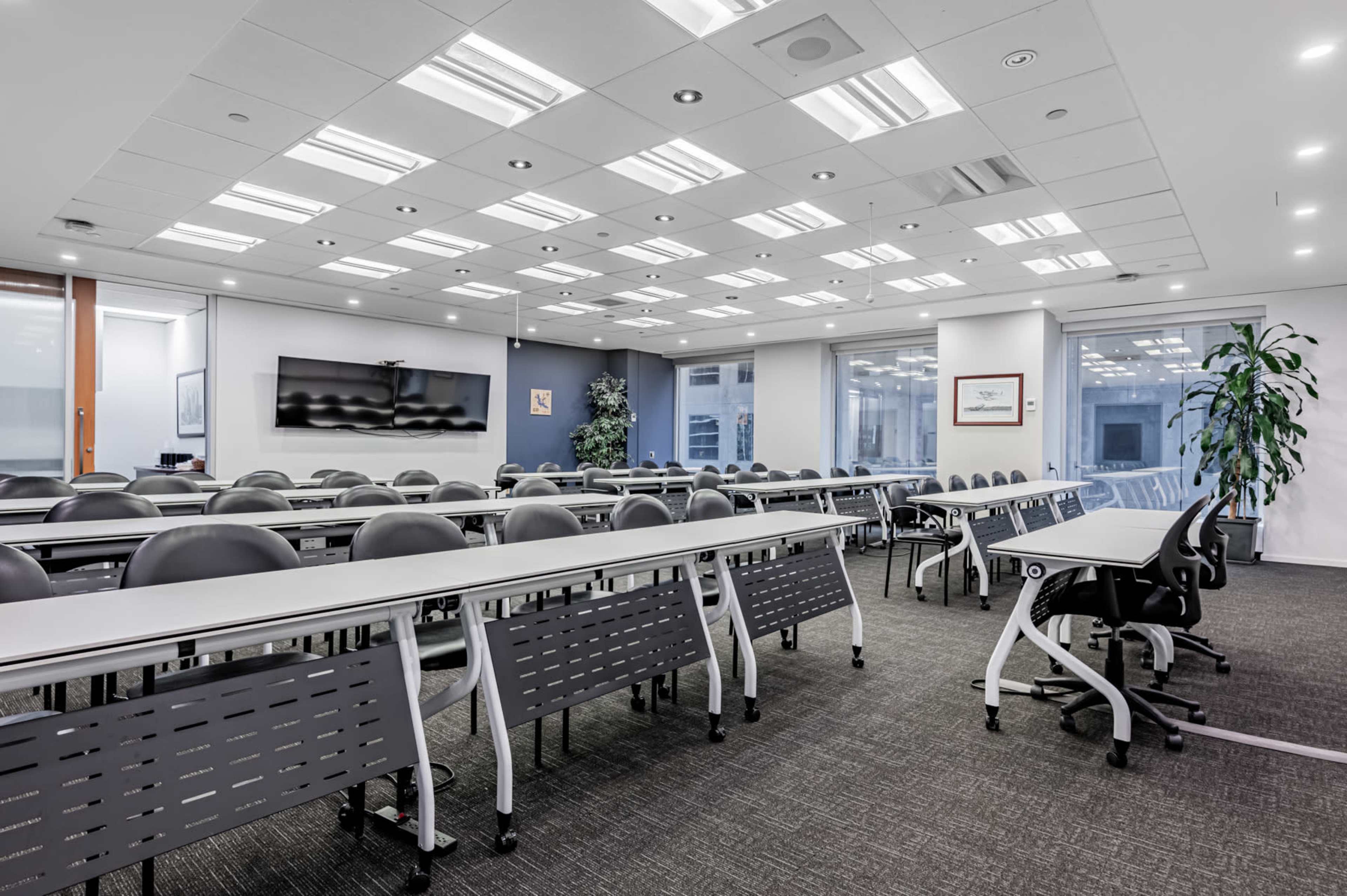 A modern conference room with rows of tables and chairs, large windows, and a wall-mounted screen.