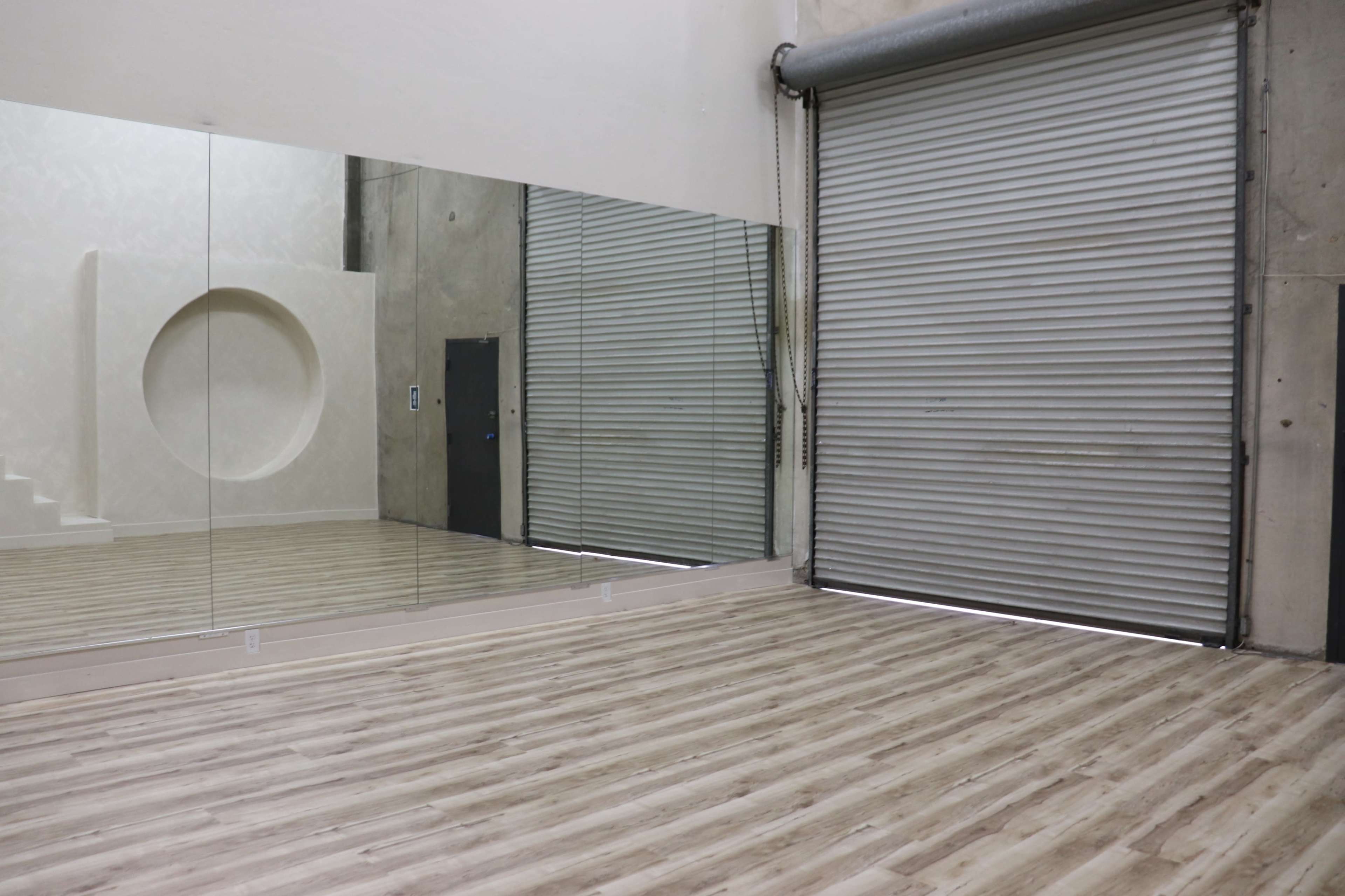 The image shows a spacious interior room with a large mirror on one wall, a closed metal roller door, and light-colored wooden flooring.