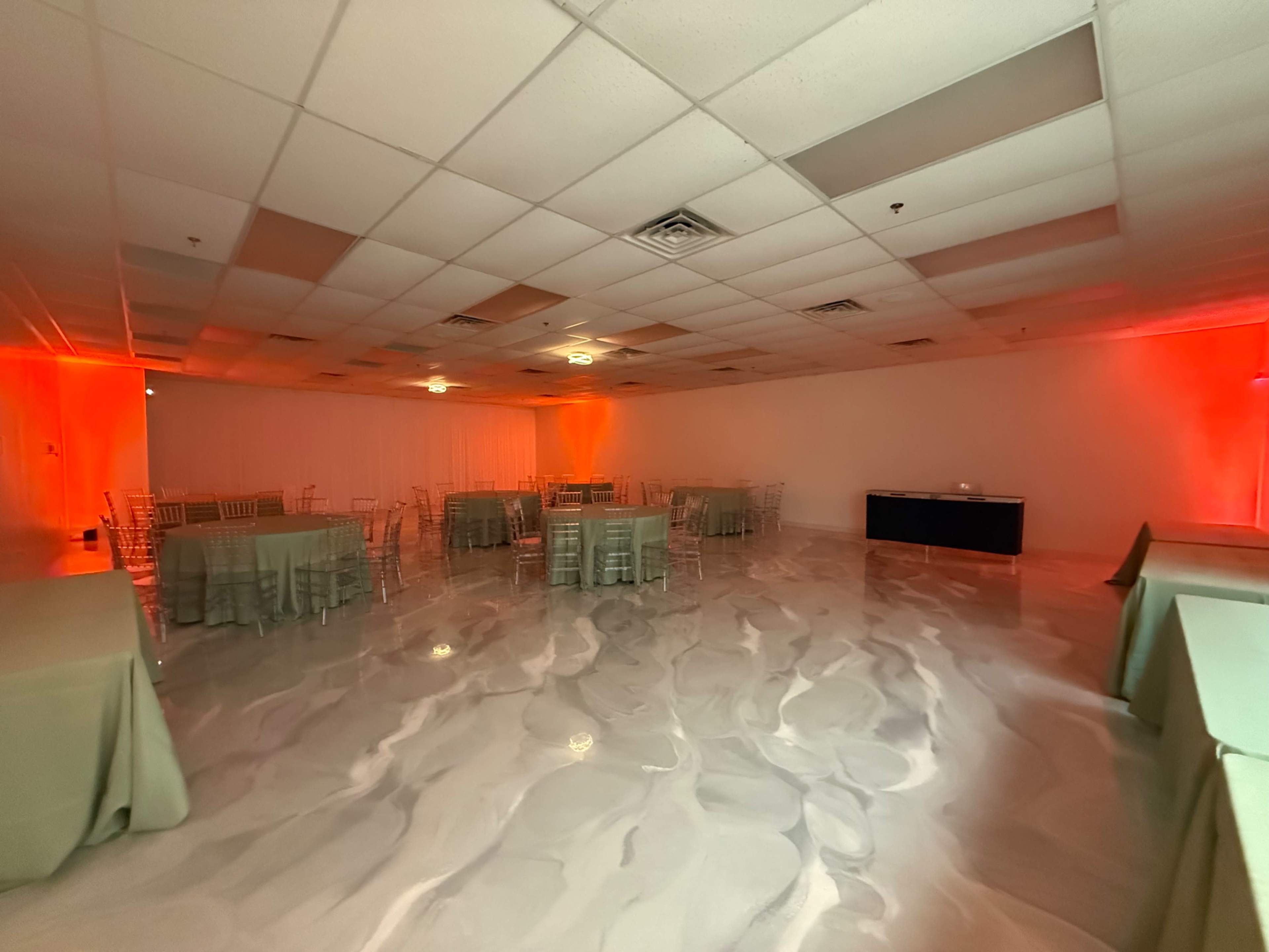 The image shows a spacious event room with round tables covered in green linens and a glossy white floor, illuminated by soft orange lighting.