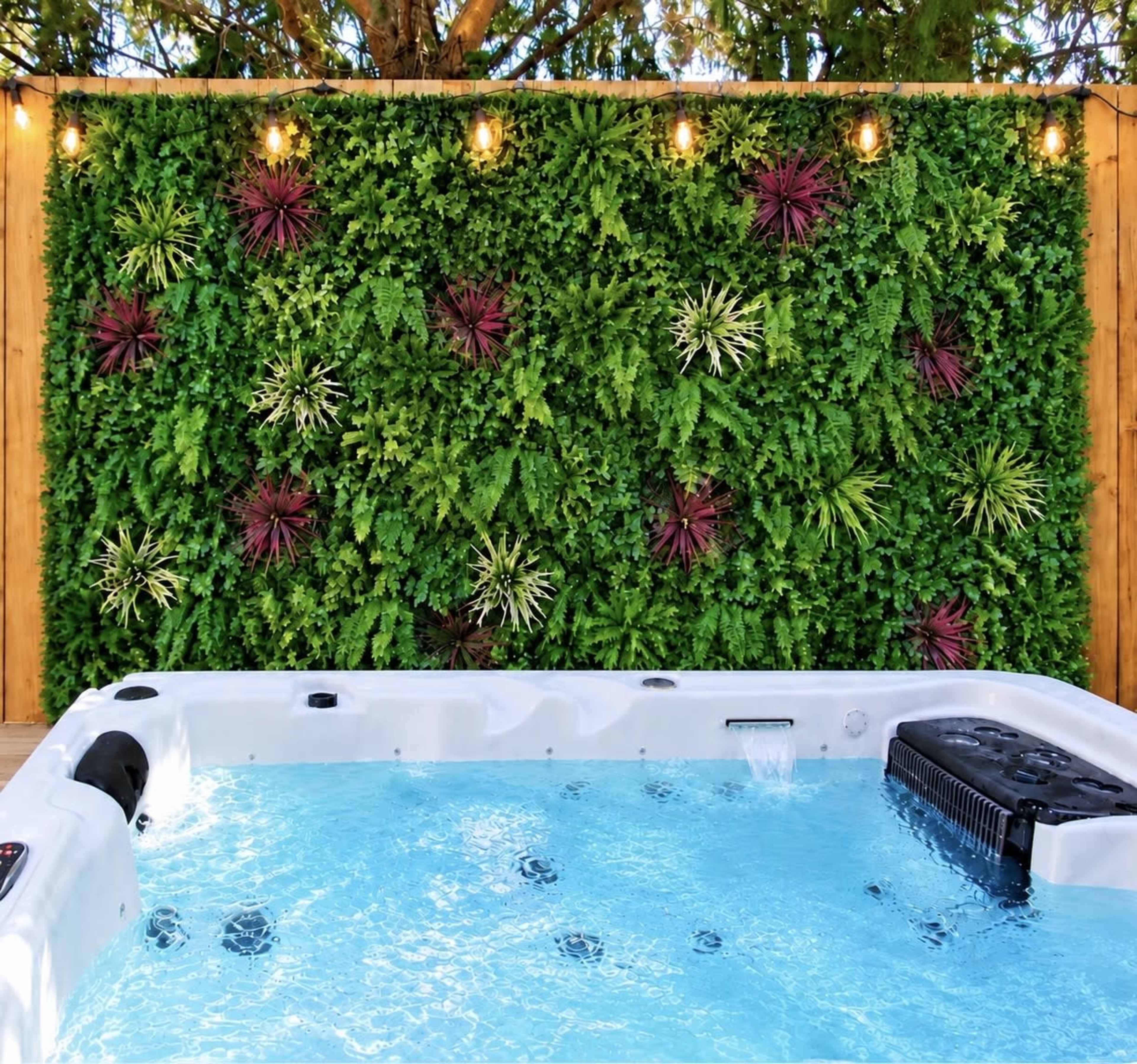 A hot tub sits in front of a wall covered with various green plants and purple accents, illuminated by string lights above.