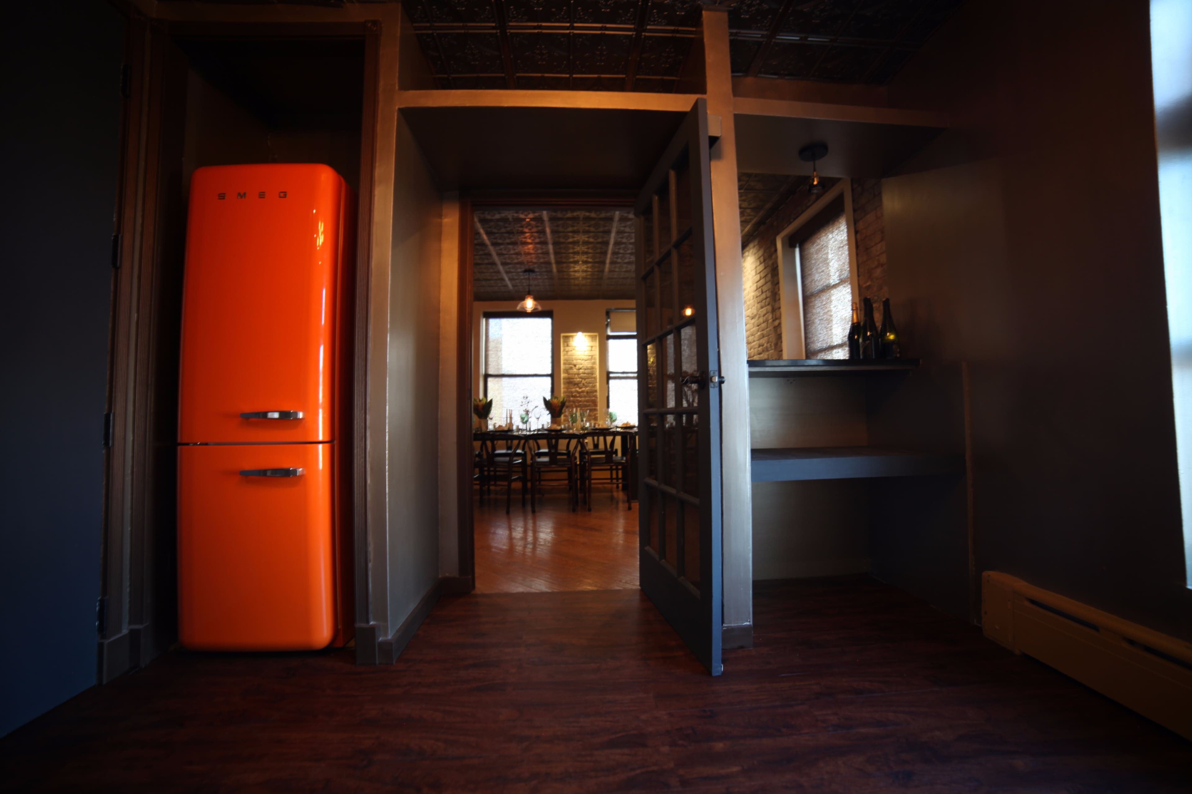 A kitchen area featuring an orange refrigerator, open double doors leading to a dining space, and wooden flooring.