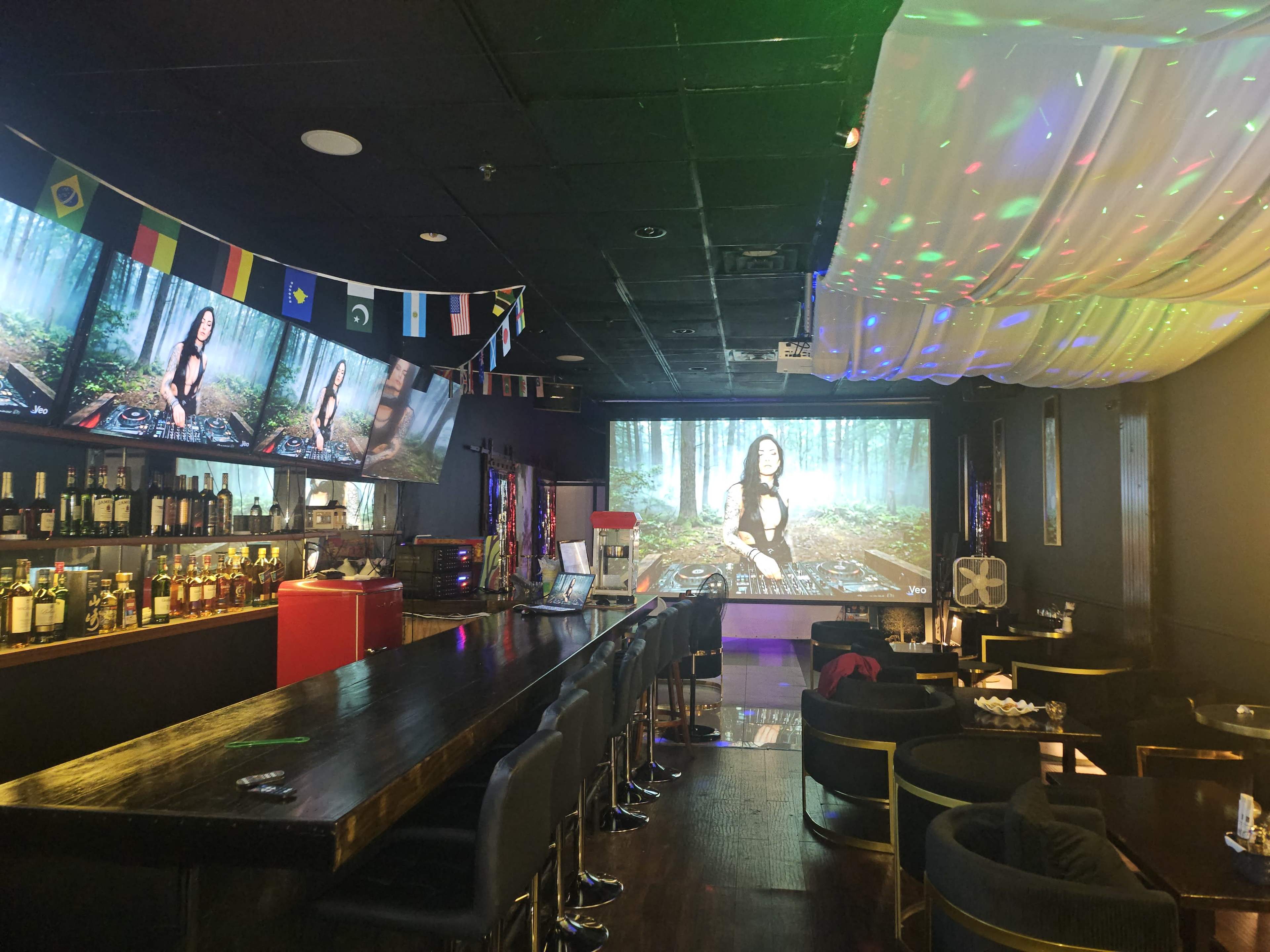 The image shows a dimly lit bar with a long wooden counter, a projection screen displaying a woman in a forest, and colorful lights reflecting from the ceiling.