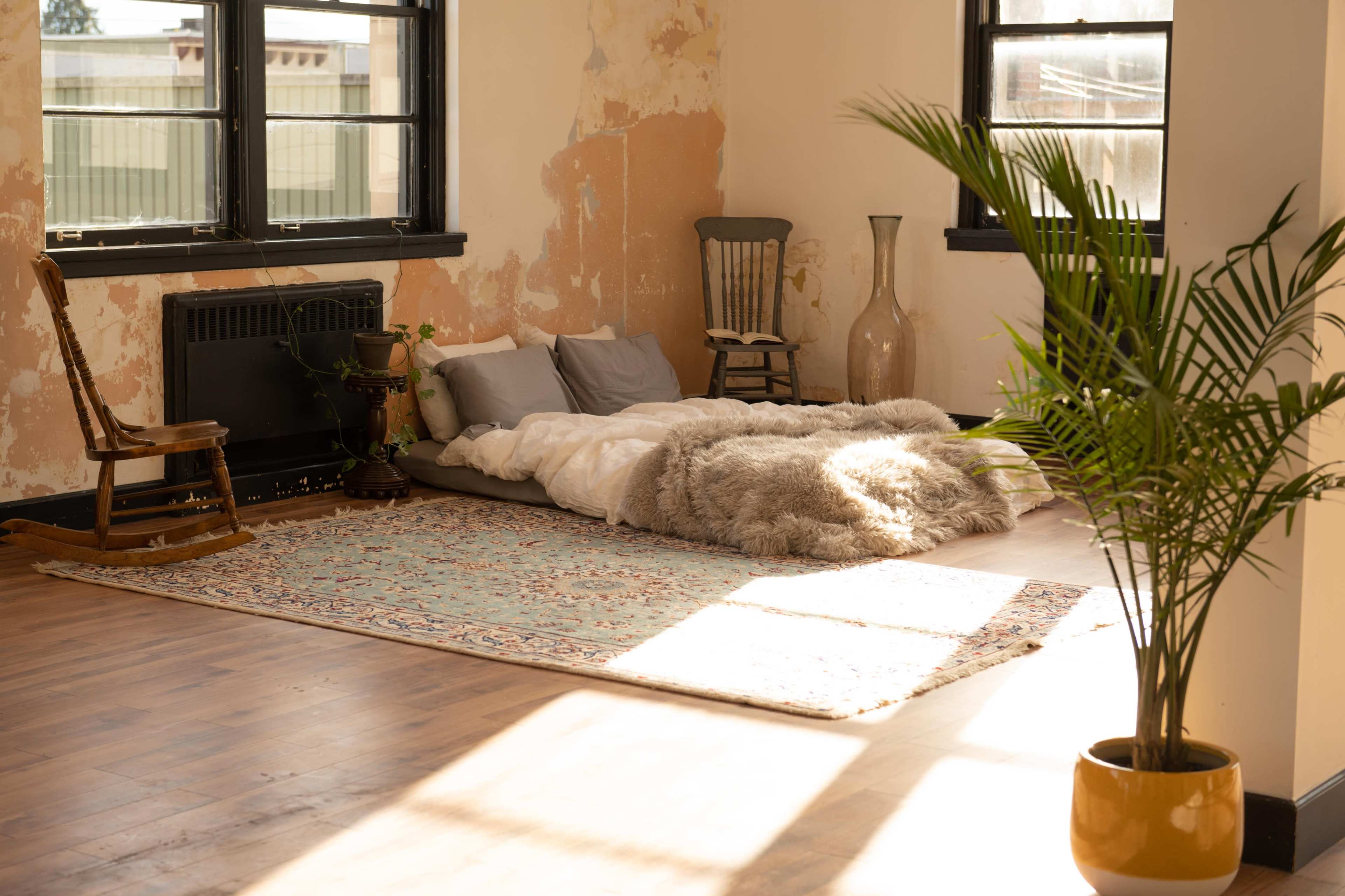 The scene features a minimalist bedroom with a bed made of blankets on a patterned rug, surrounded by chairs and a potted plant in a sunlit, partially distressed room.