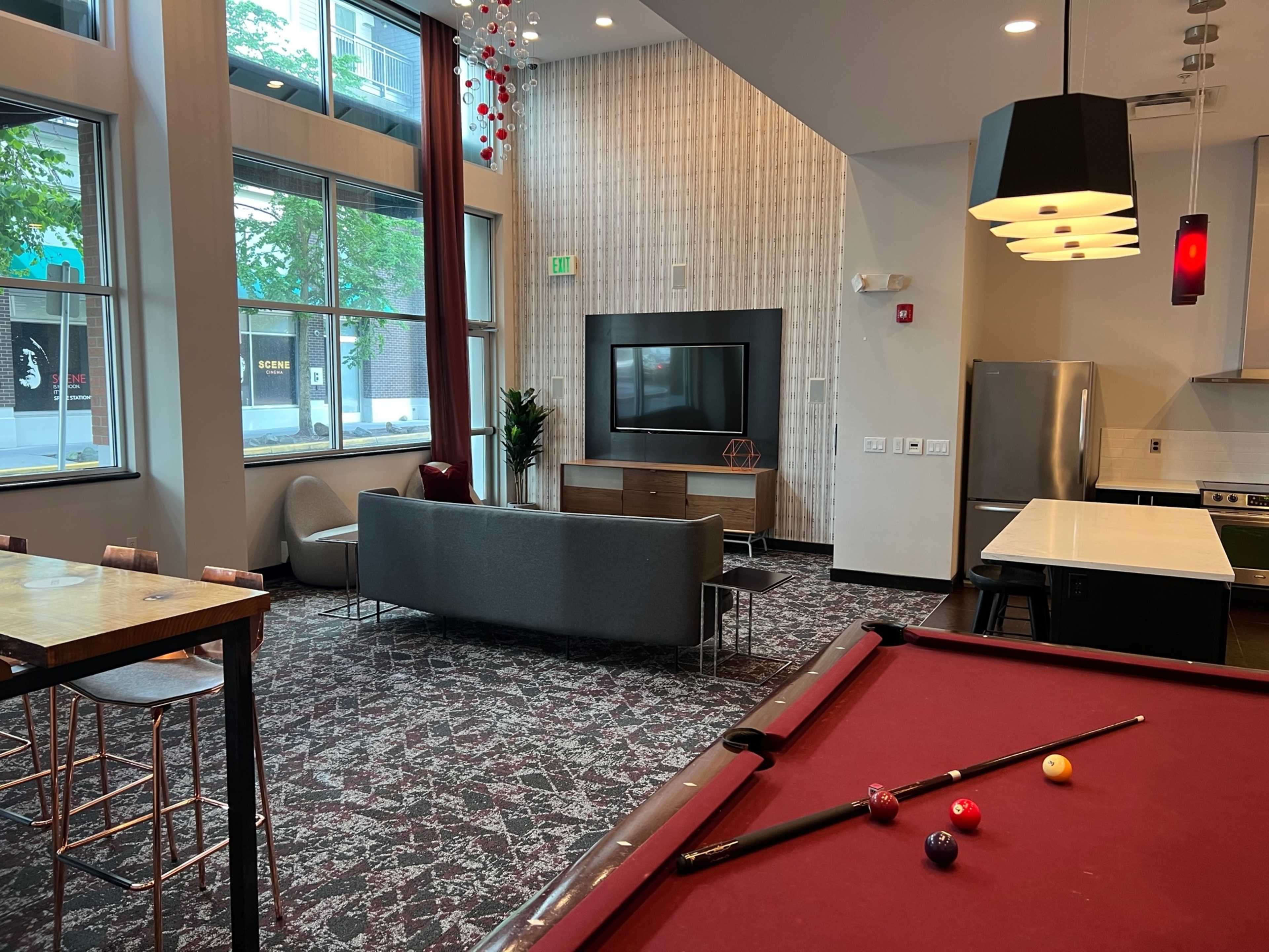 A lounge area features a pool table in the foreground, a modern seating arrangement, and large windows letting in natural light.