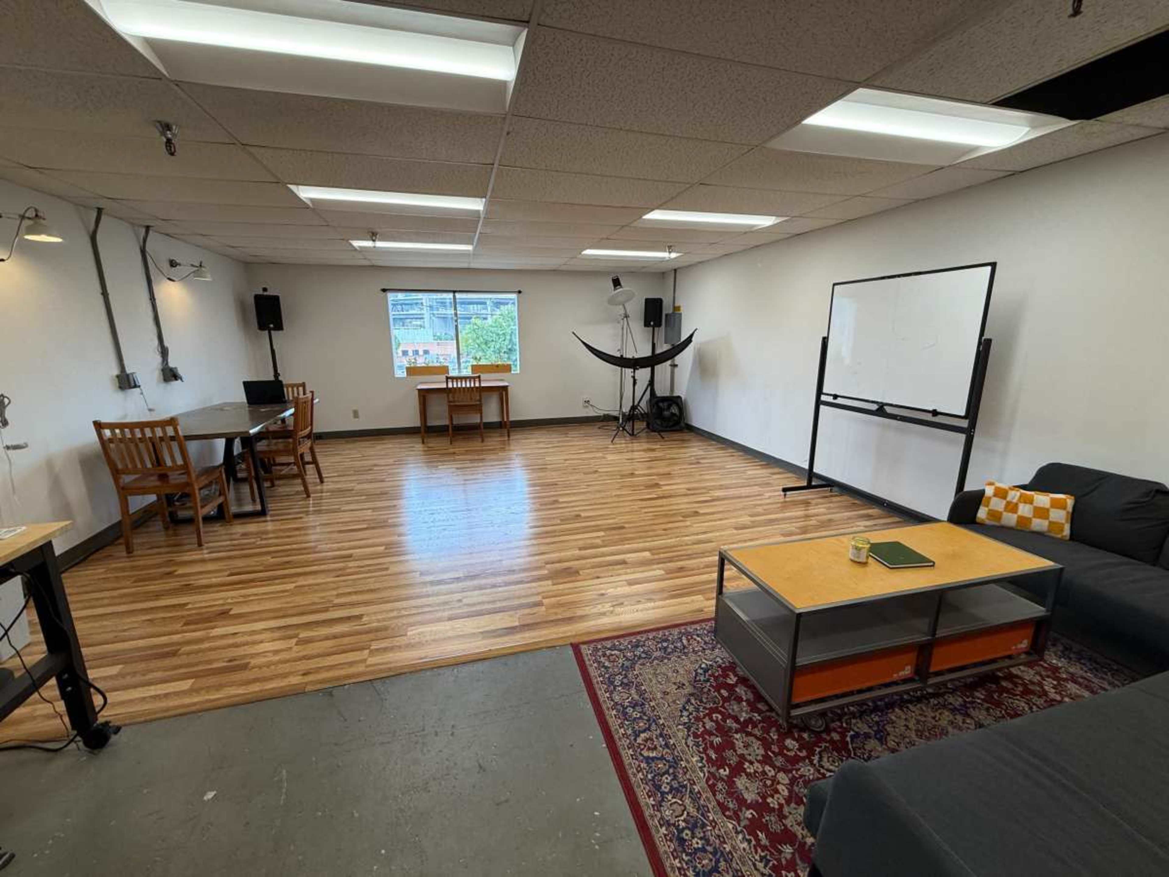 The image shows a spacious room with wooden flooring that includes a sofa, a coffee table, a dining table with chairs, a whiteboard, and studio lighting equipment.