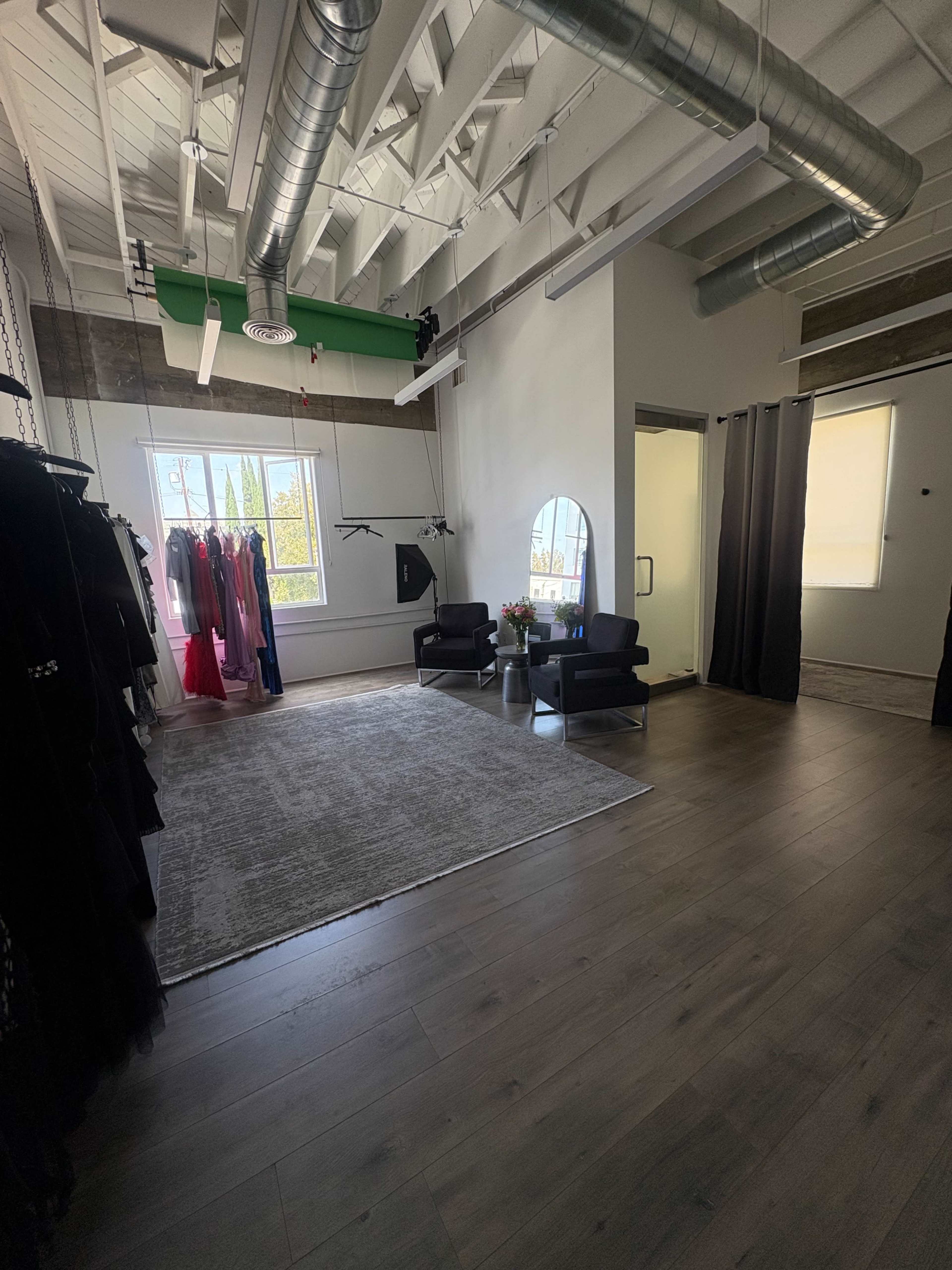 The image shows a spacious, well-lit room featuring elegant black chairs, a gray rug, and a rack of dresses, along with large windows allowing natural light to enter.