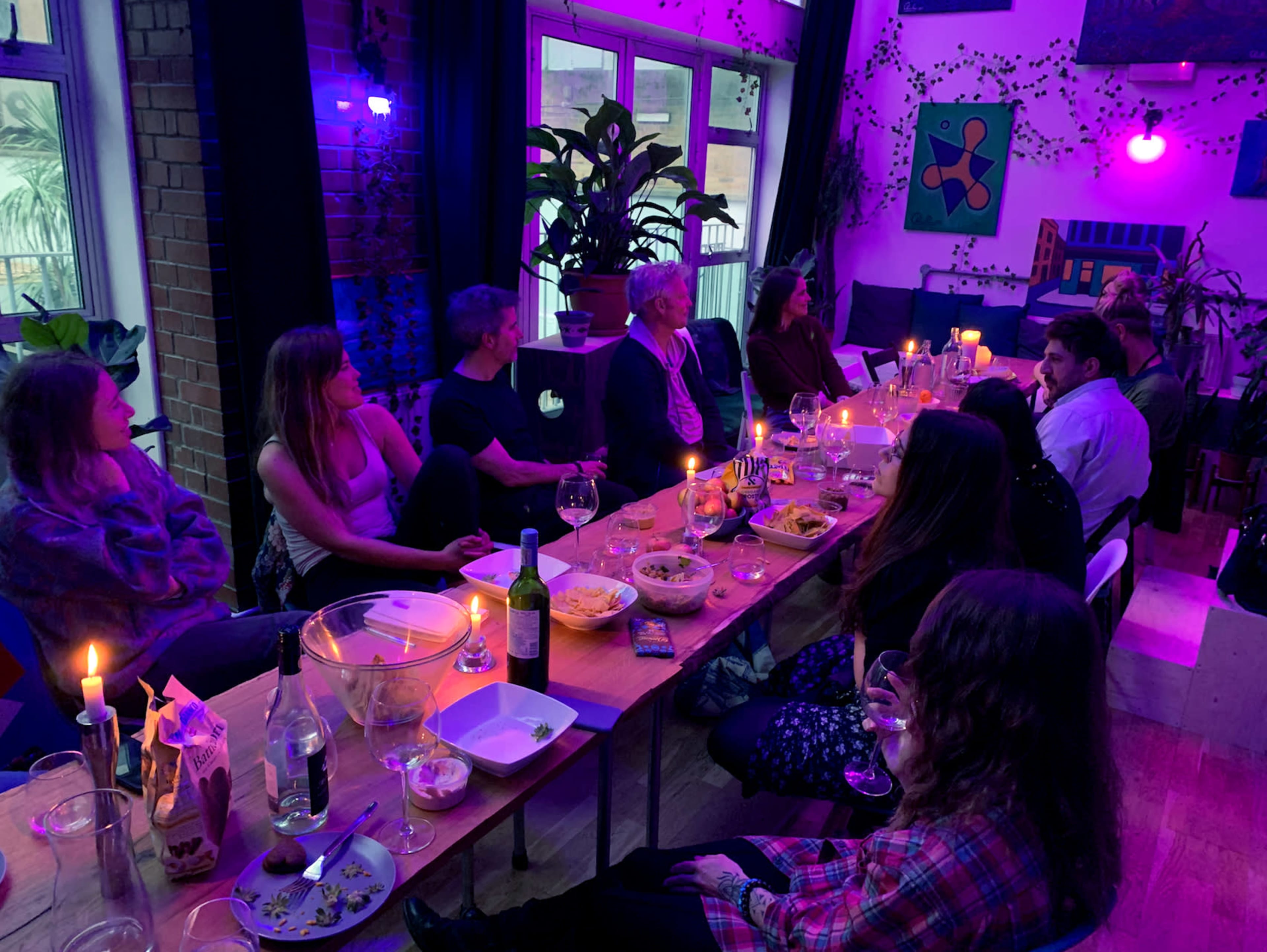 A group of people sits around a long table decorated with food and drinks in a dimly lit room filled with plants and colorful artwork.