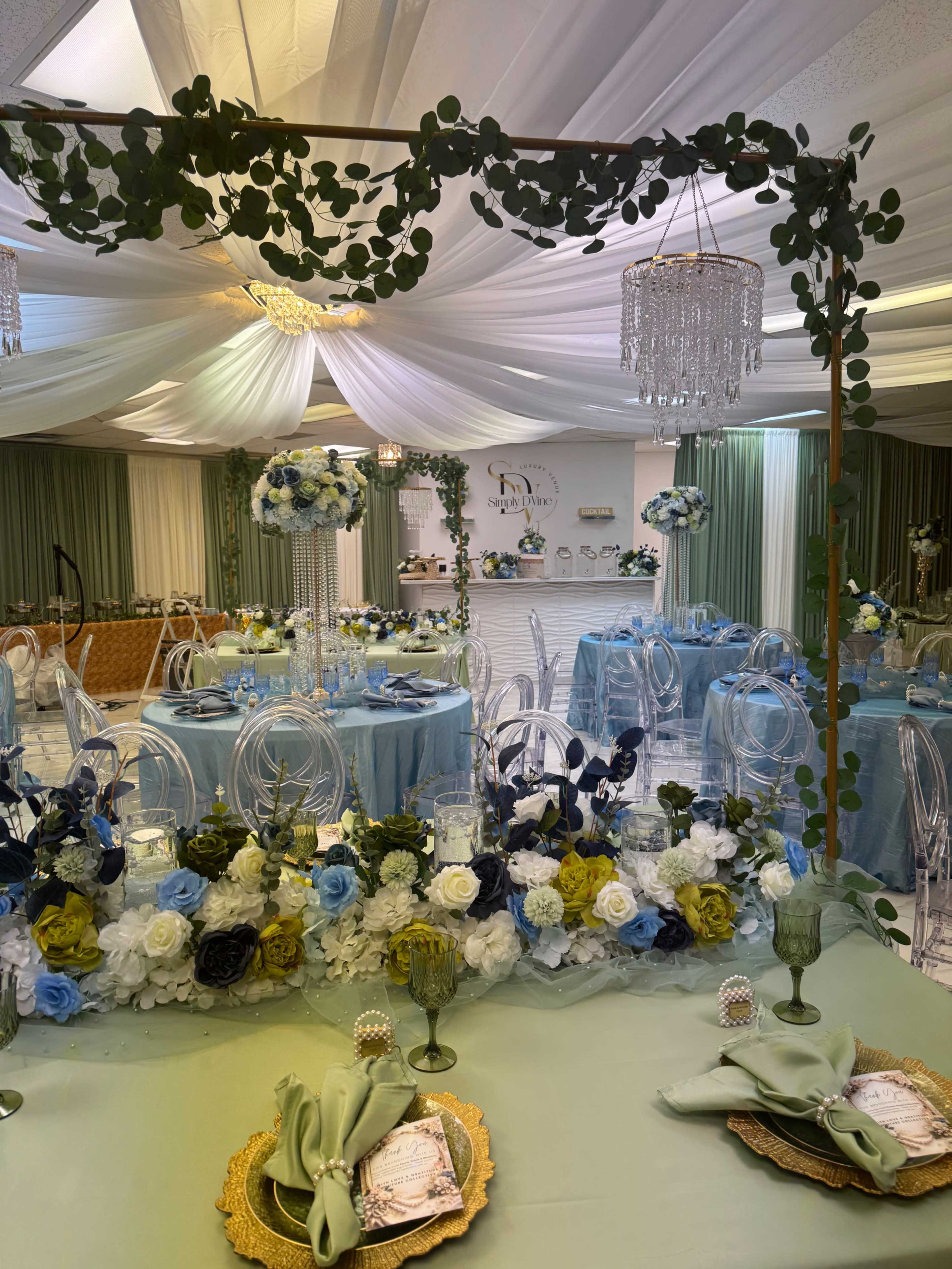 The image shows a decorated event space with elegantly set tables featuring floral arrangements, tablecloths in shades of blue and green, and chandeliers hanging from a draped ceiling.