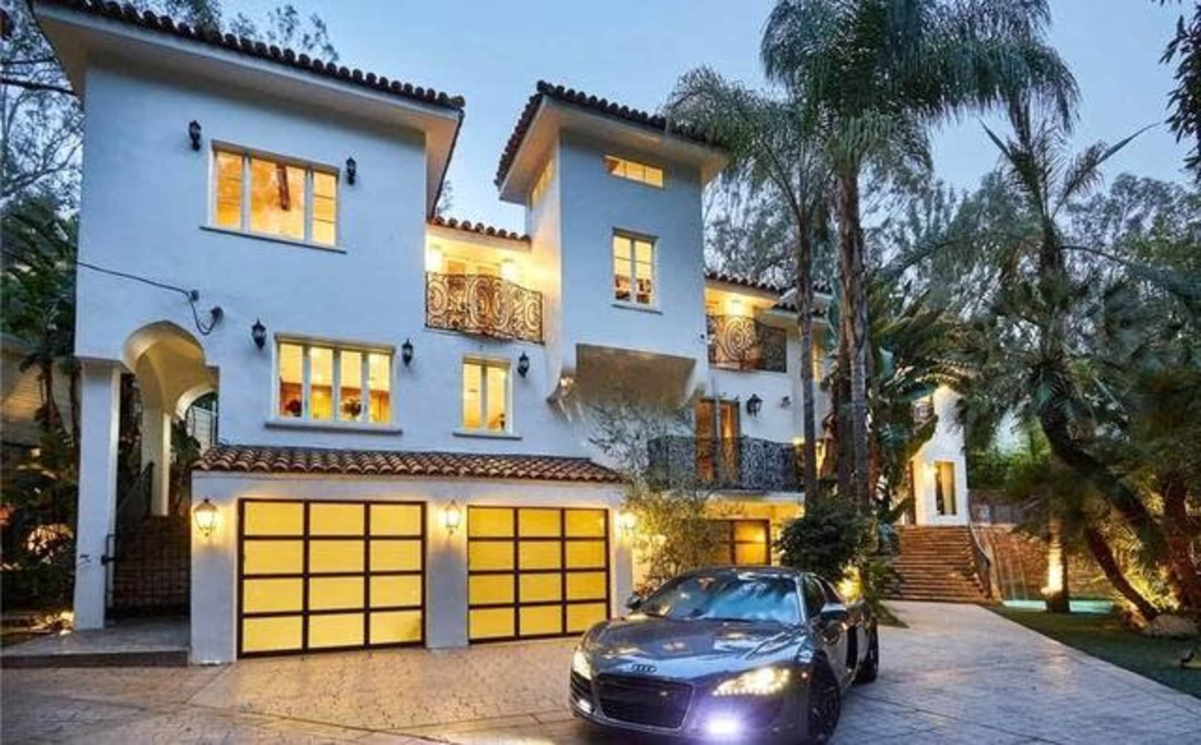 The image shows a two-story white stucco house with a tile roof, featuring multiple balconies and a landscaped driveway with a parked black sports car.