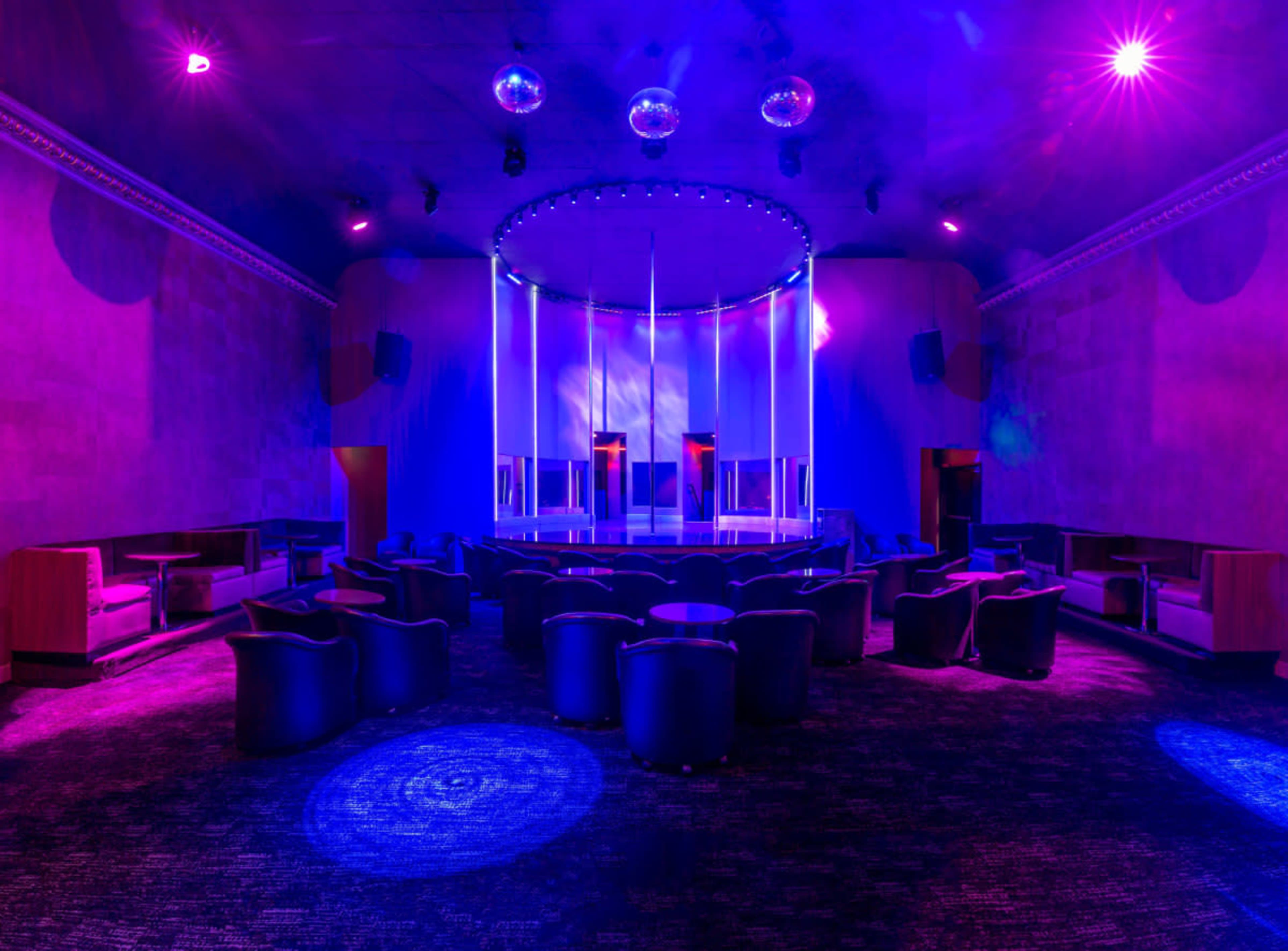 A dimly lit nightclub interior with a central stage, surrounding lounge seating, and multiple disco balls hanging from the ceiling.