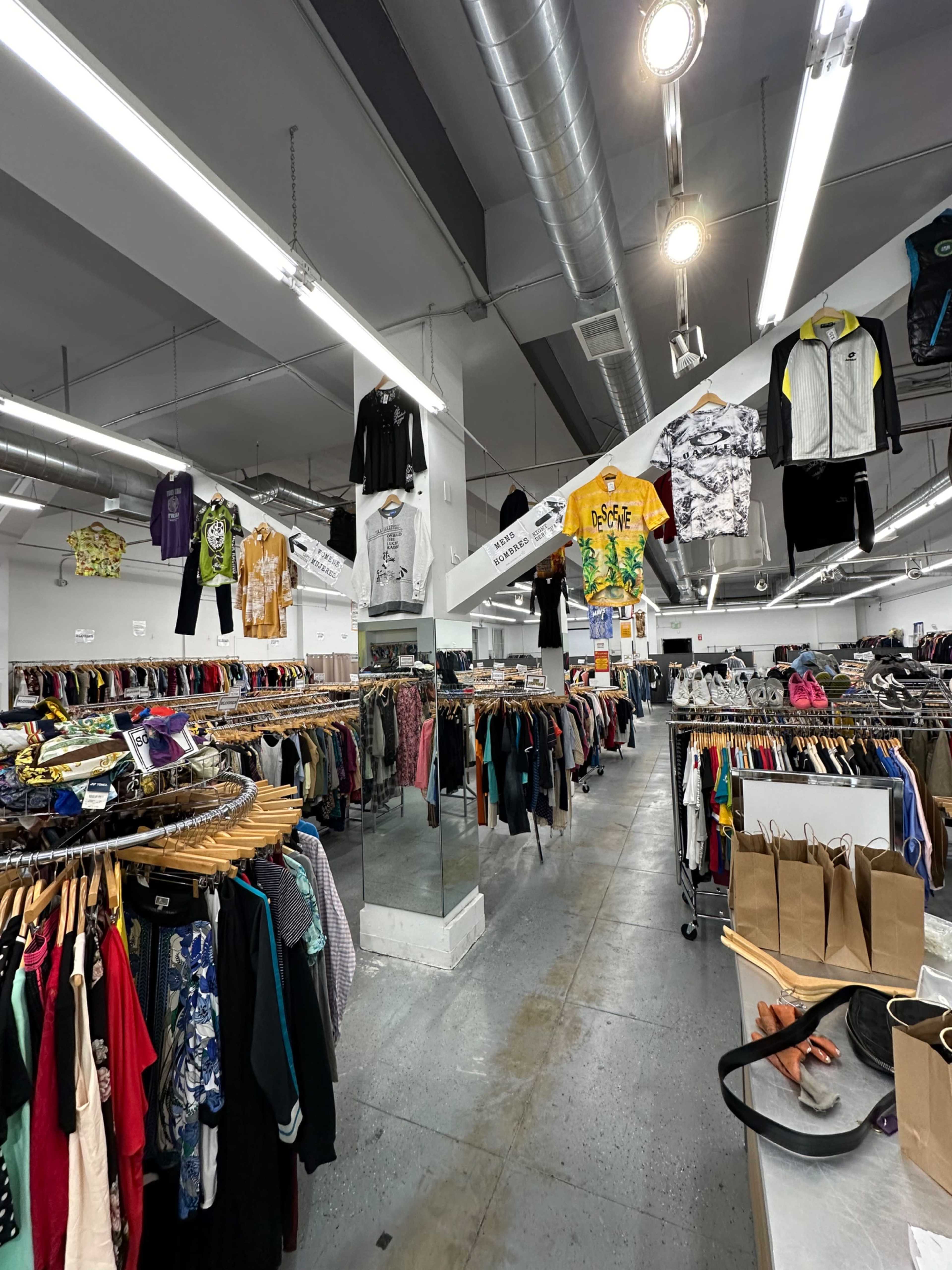 The image shows the interior of a clothing store filled with racks of garments, including hanging items from the ceiling and a mirror along one wall.