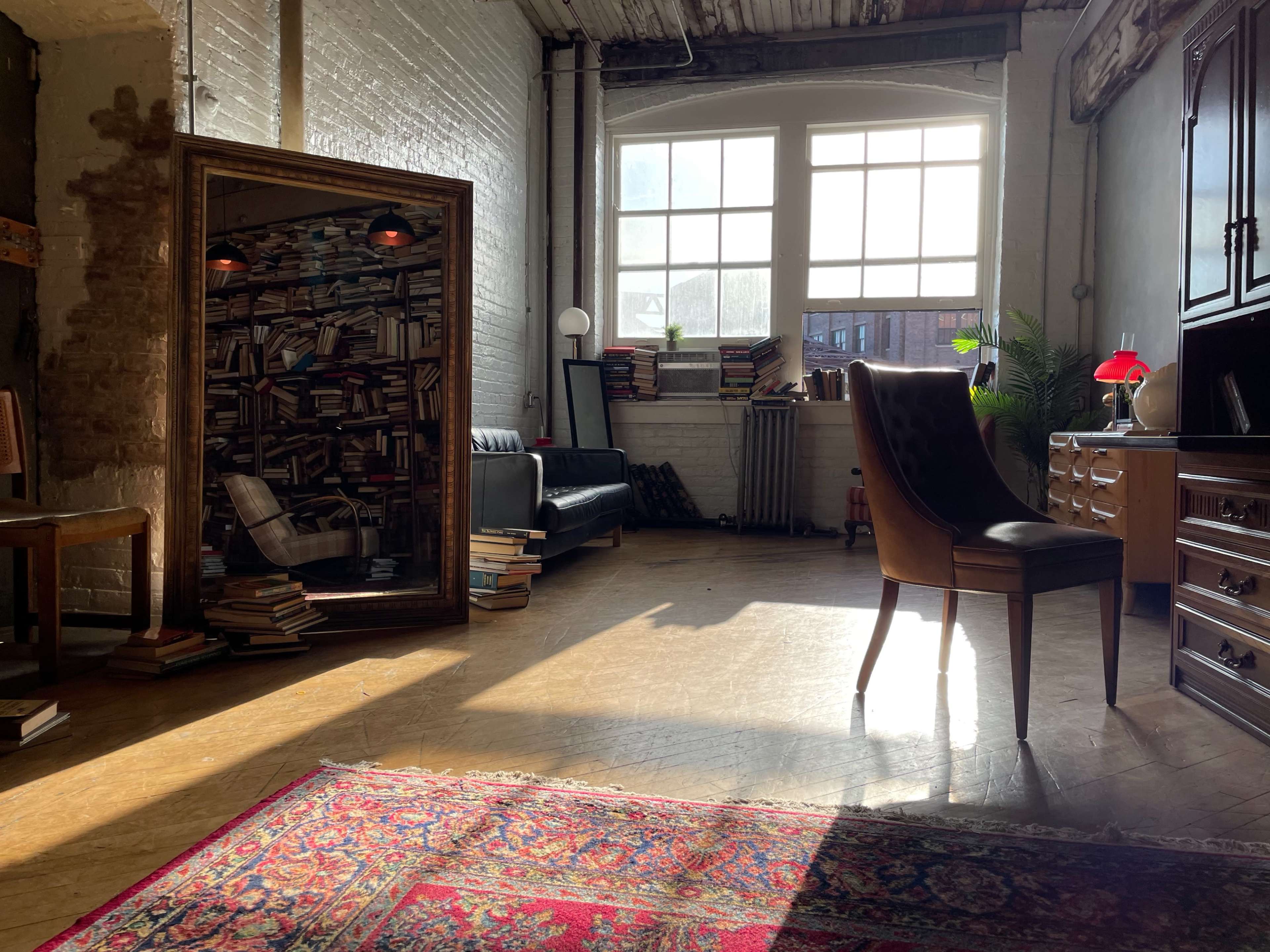 A sunlit room features a large mirror with a bookshelf design, a leather couch, a wooden desk with a chair, and a colorful rug on the floor.
