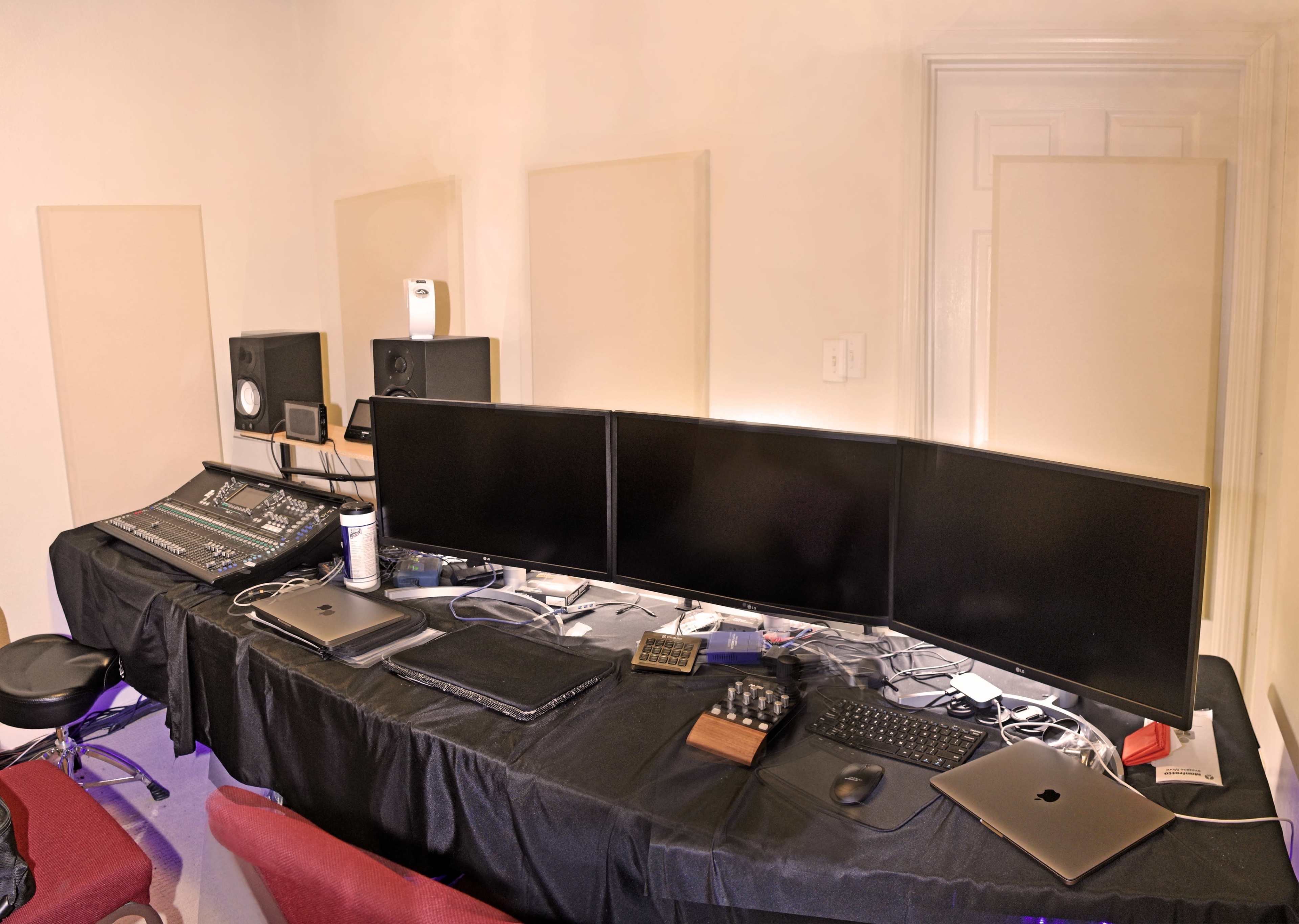 A workstation is set up with multiple monitors, a mixing console, laptops, and audio equipment arranged on a black table.
