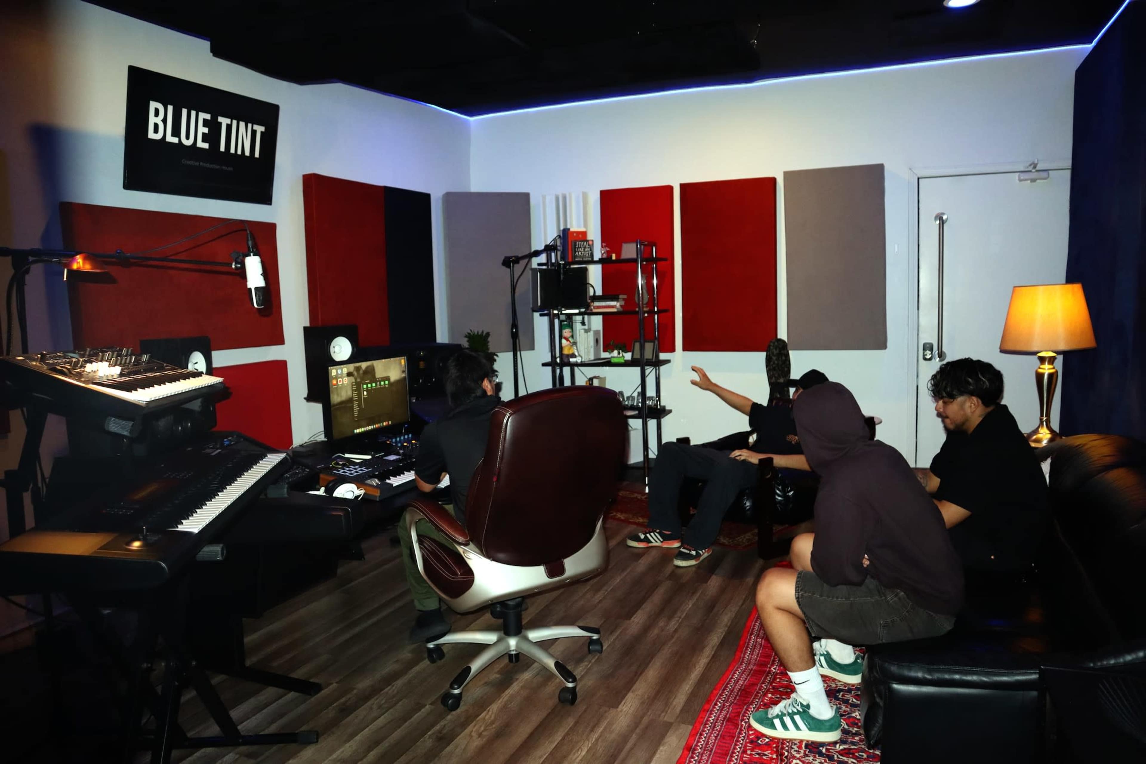 The image shows a music studio with a keyboard, mixing equipment, and four people engaged in conversation while sitting on a couch and a chair.