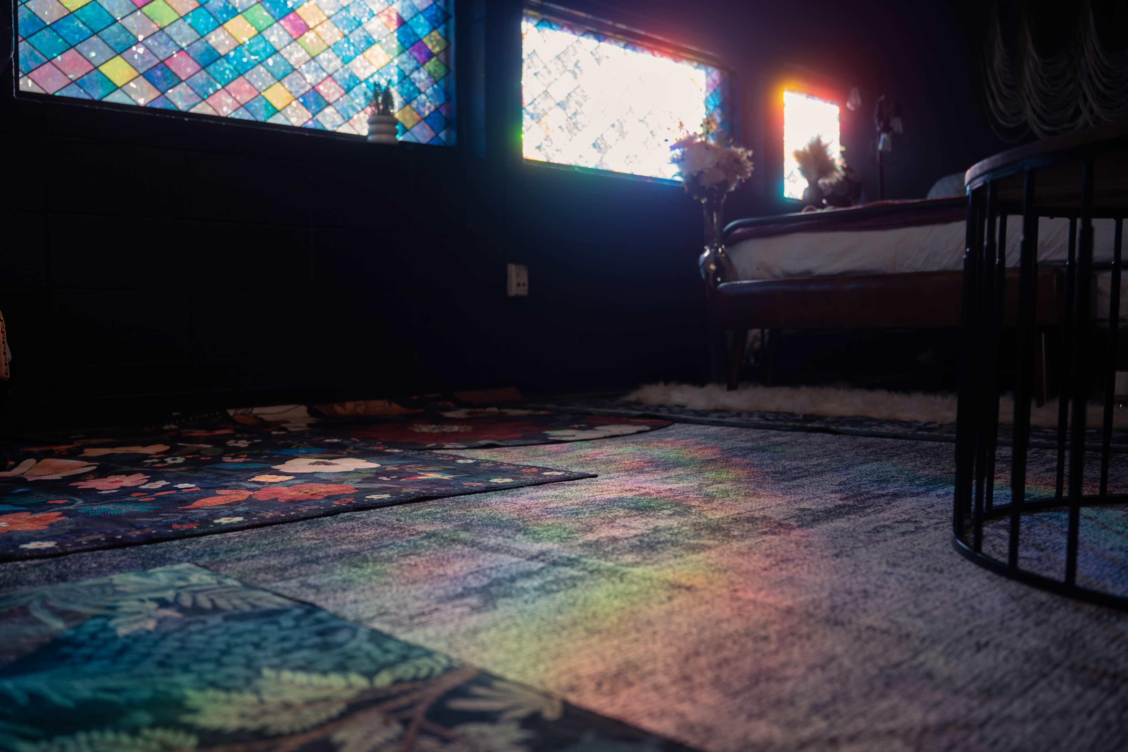 The image shows a room with colorful stained glass windows casting light onto patterned rugs and a bed in the background.