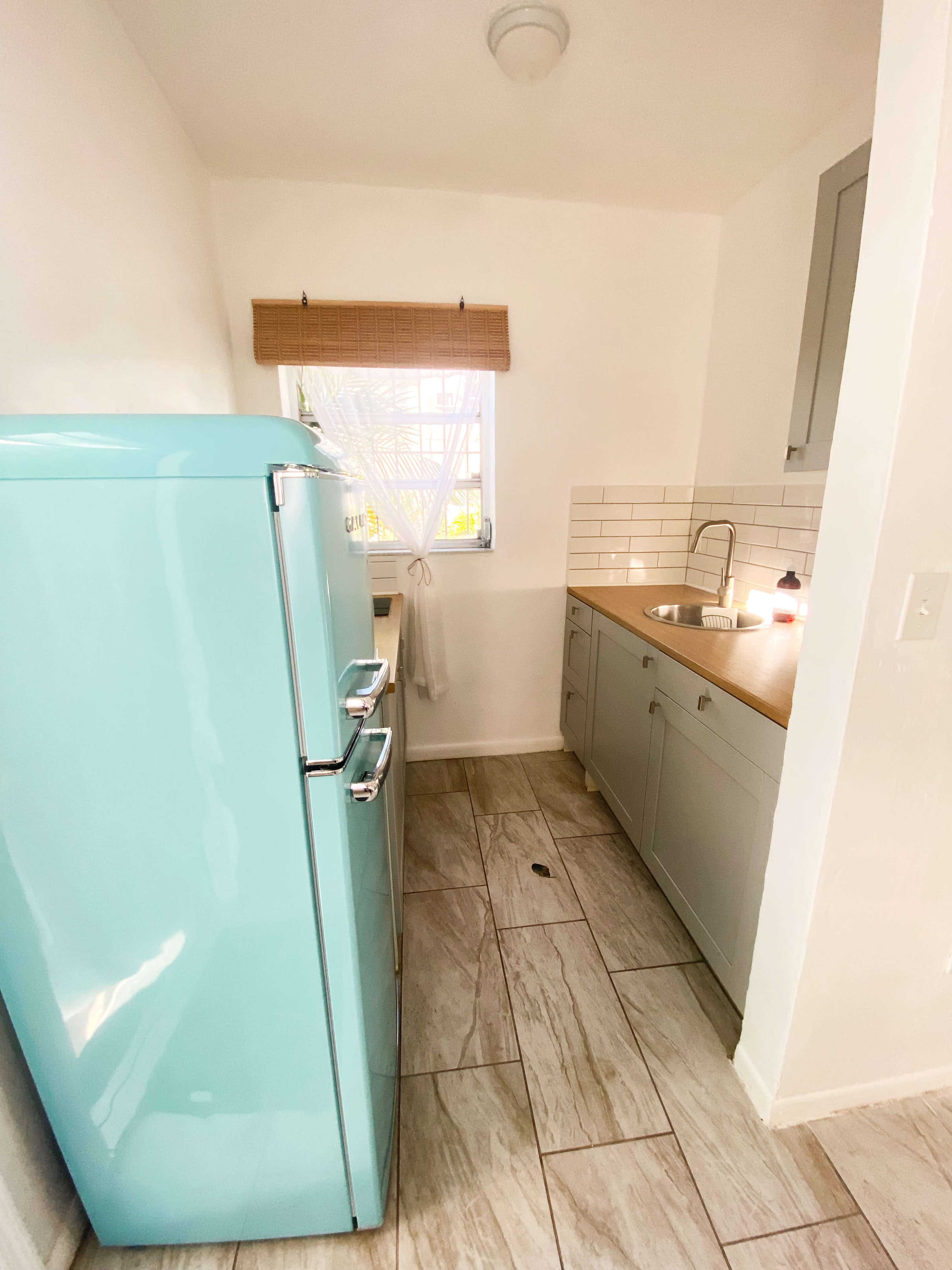 A light blue vintage refrigerator stands next to a modern kitchen sink and countertop with white tiles in a small, brightly lit kitchen space.