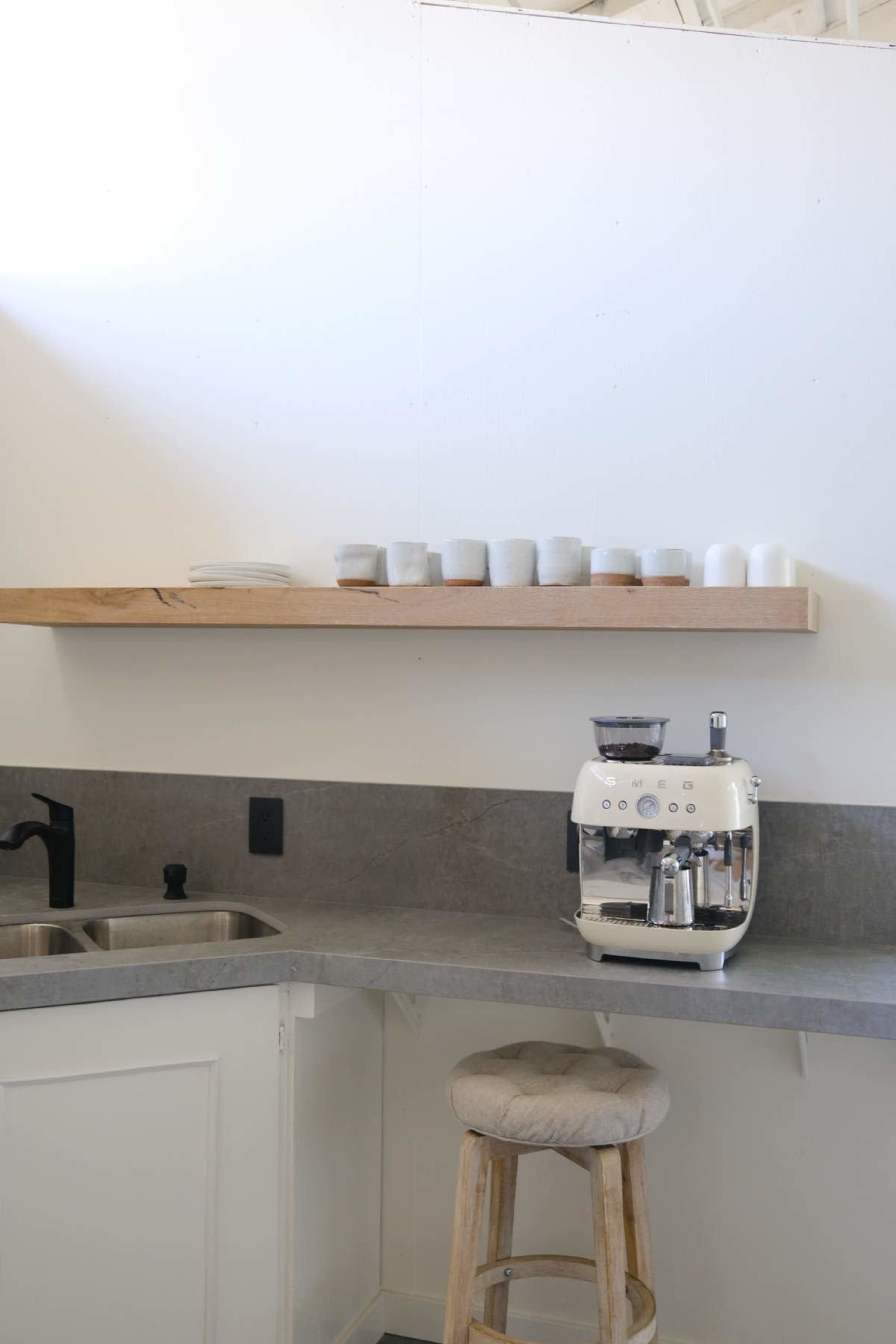 The image shows a minimalist kitchen corner featuring a coffee machine on a gray countertop, with a wooden shelf above displaying several cups.