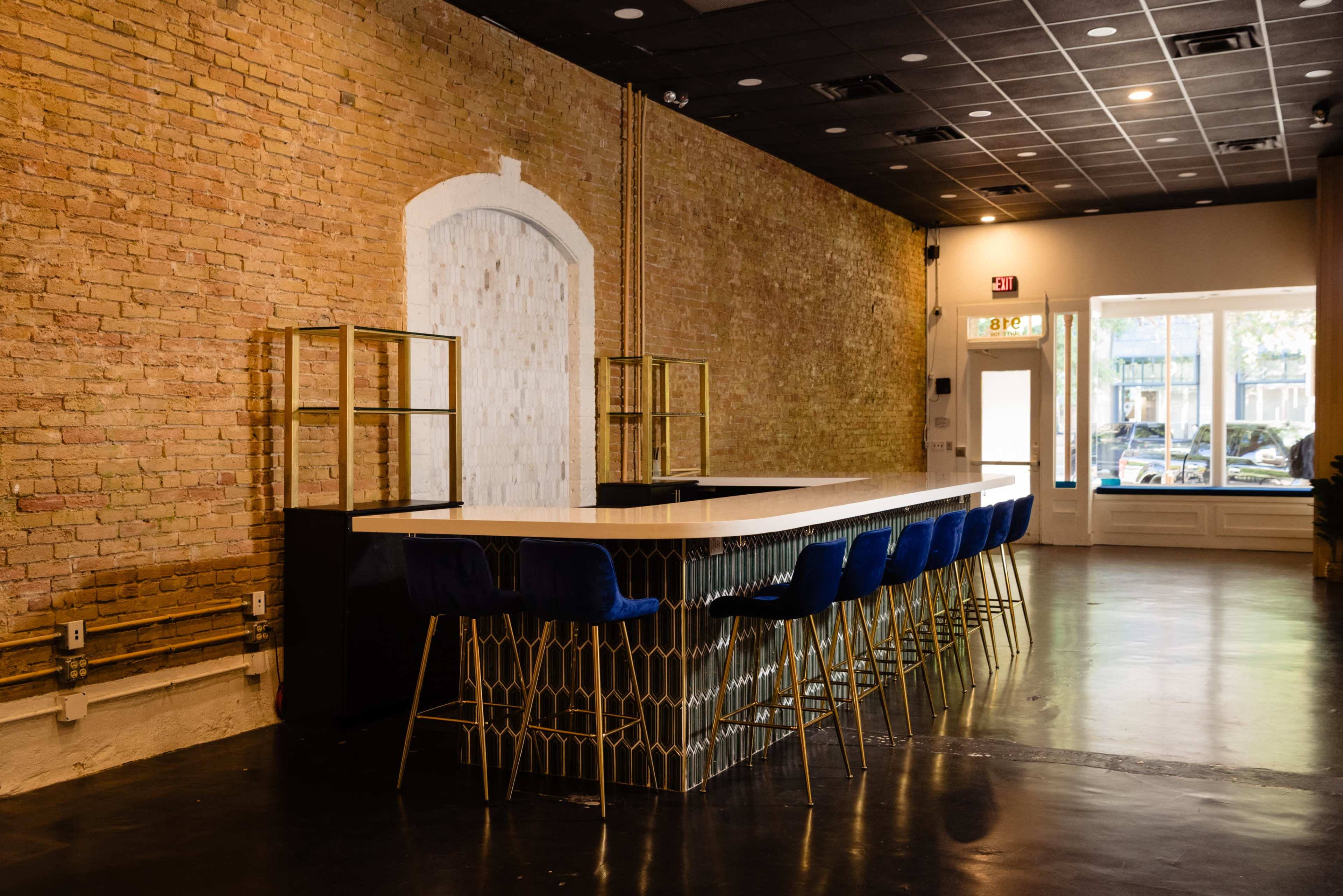 The image shows a modern bar with a white countertop, blue chairs, and an exposed brick wall in a well-lit interior space.