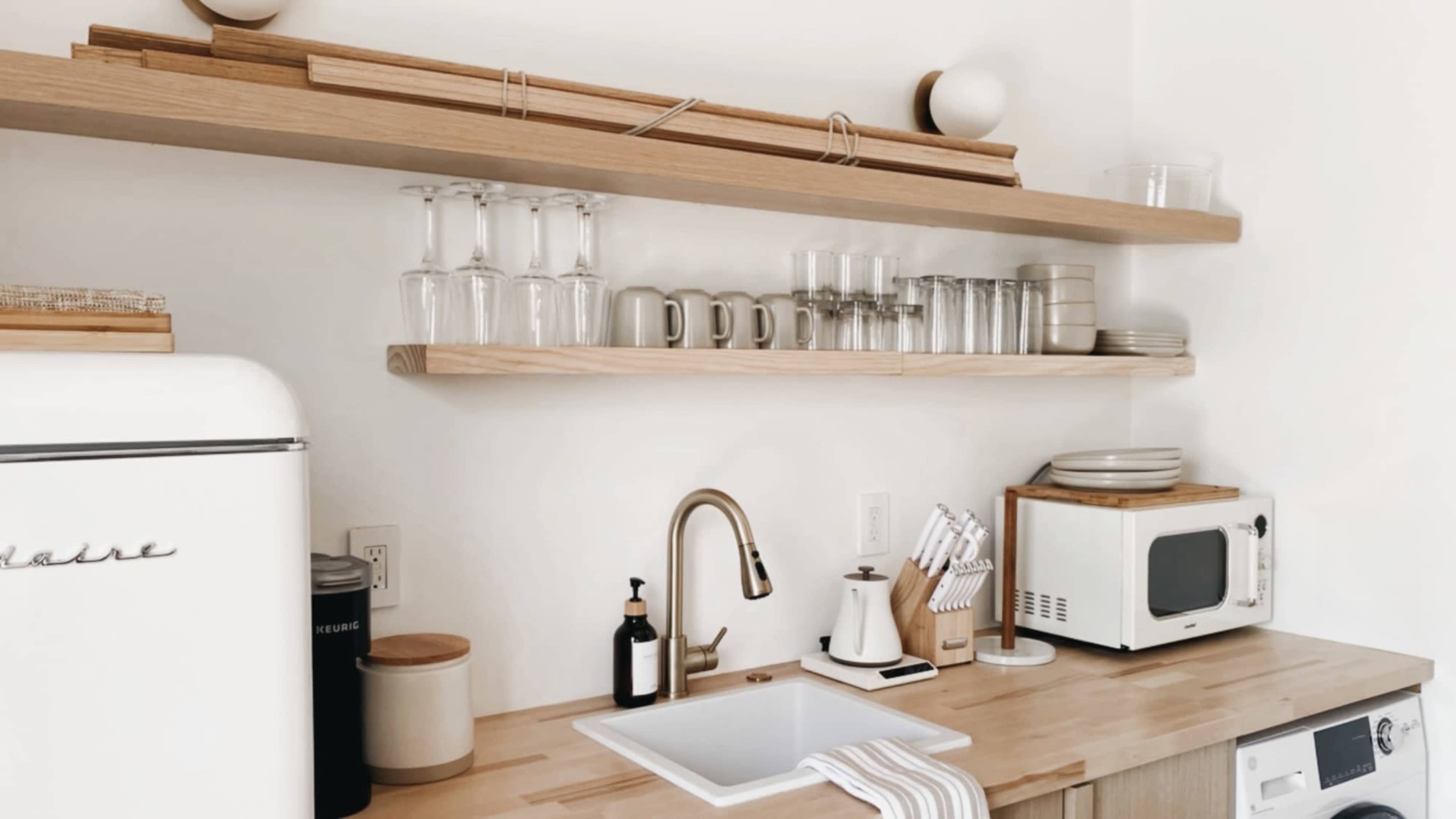 The image shows a modern kitchen with wooden shelves holding glassware and dishes, a stainless steel sink, and a microwave on the counter.