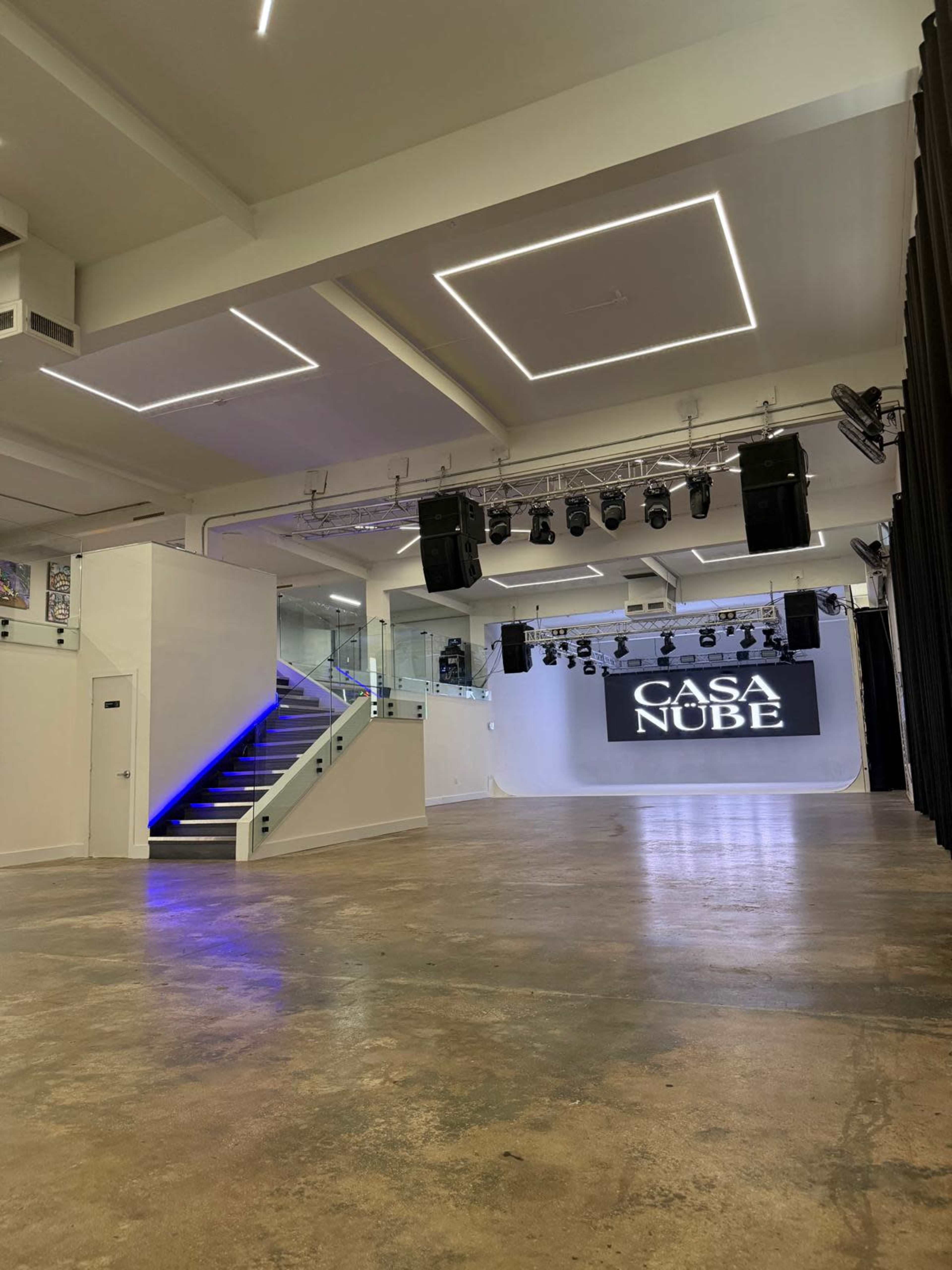The image shows a spacious interior of a venue featuring a stage with lighting, sound equipment, and a sign that reads "CASA NUBE."