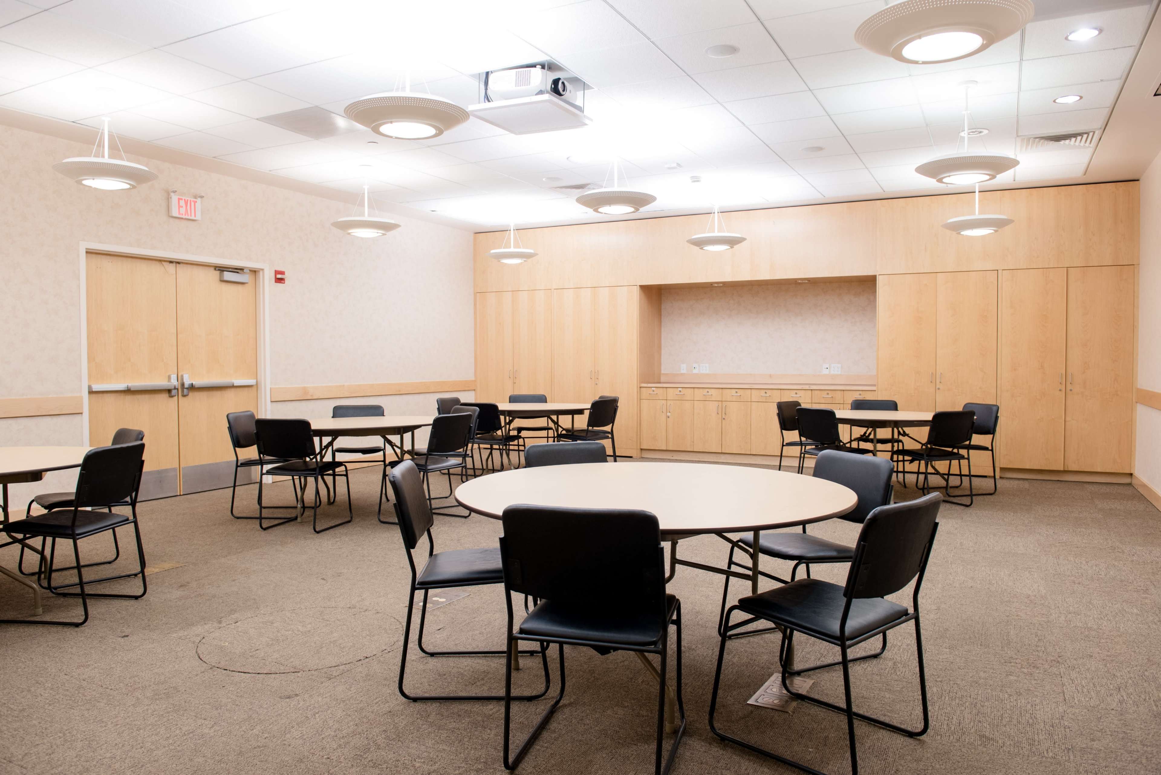 A spacious meeting room with several round tables and chairs arranged on a carpeted floor, illuminated by overhead lights.