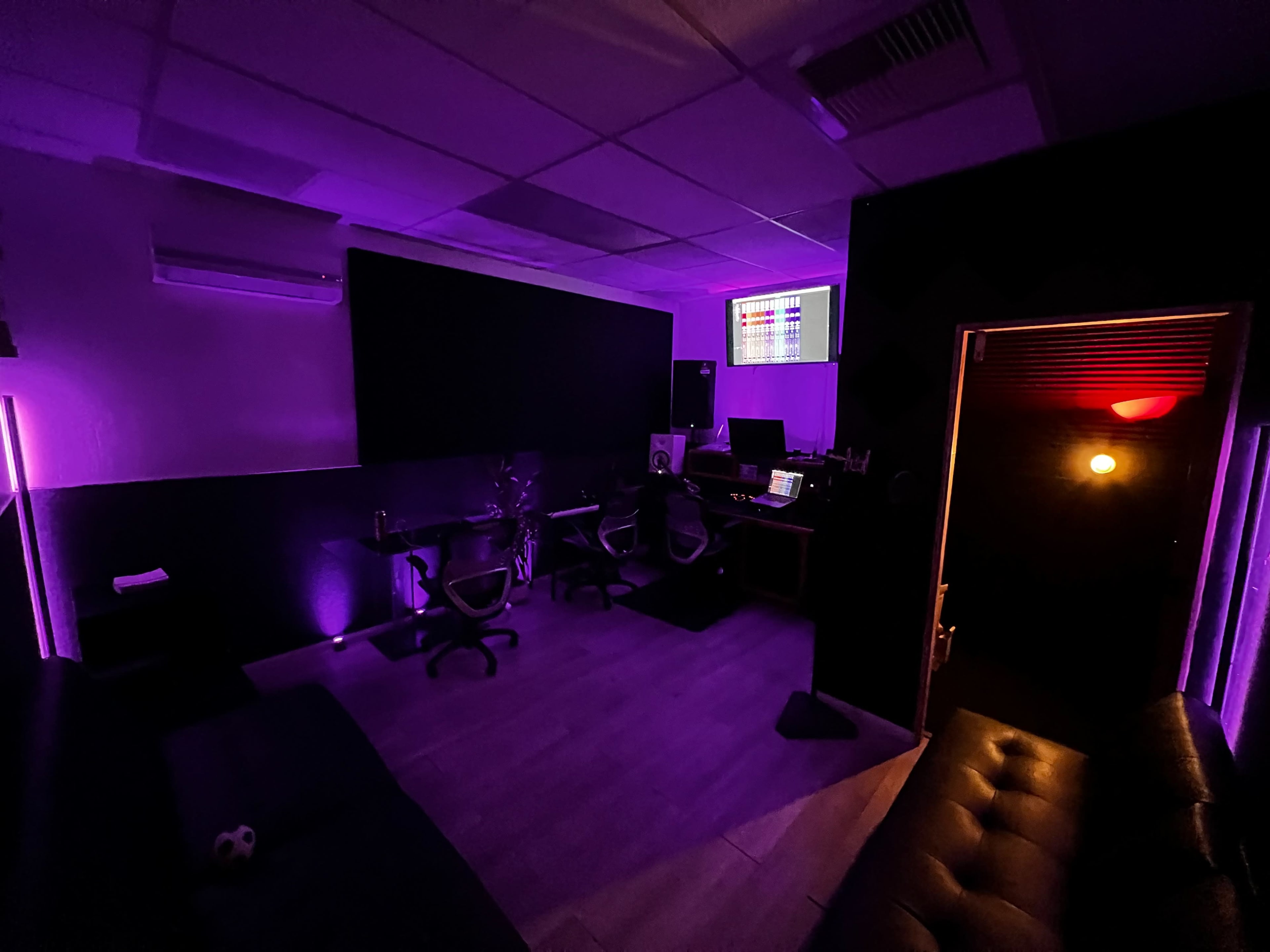 A dimly lit recording studio features purple lighting, a large black wall, and workstations with chairs.