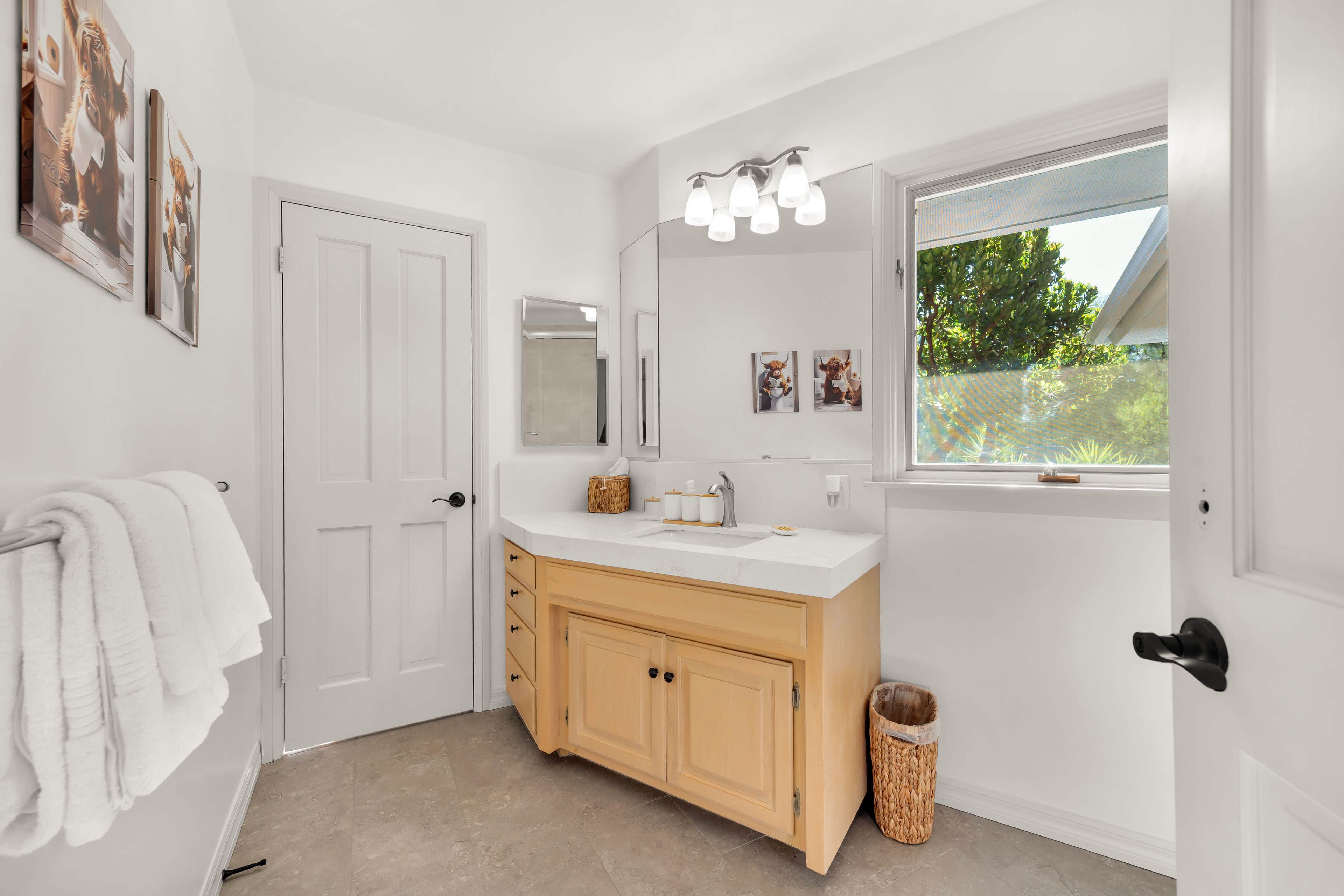 The image shows a well-lit bathroom featuring a light-colored vanity with a marble countertop, a mirror, and a window overlooking greenery.