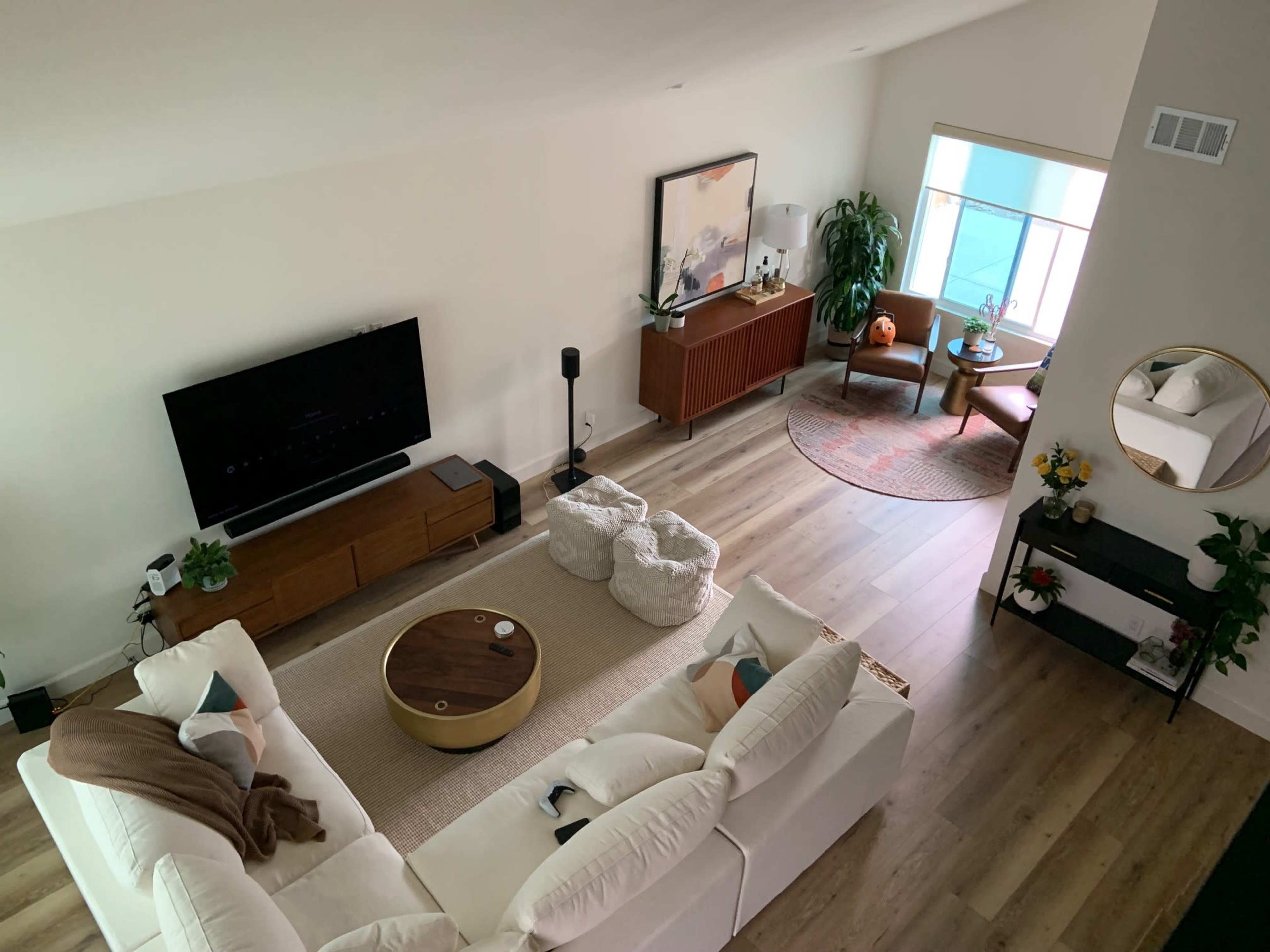 A spacious living room features a white sectional sofa, a circular coffee table, a television on a wooden stand, and a sunlit corner with a chair and a plant.