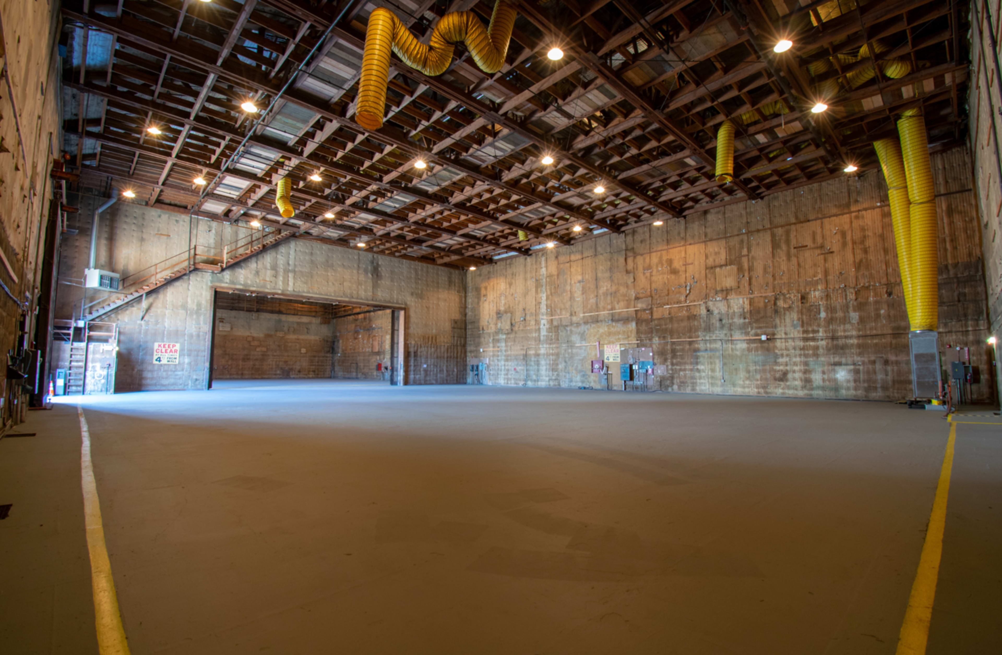 The image shows a large, empty warehouse interior featuring high ceilings, exposed beams, and yellow ventilation ducts hanging from the ceiling.