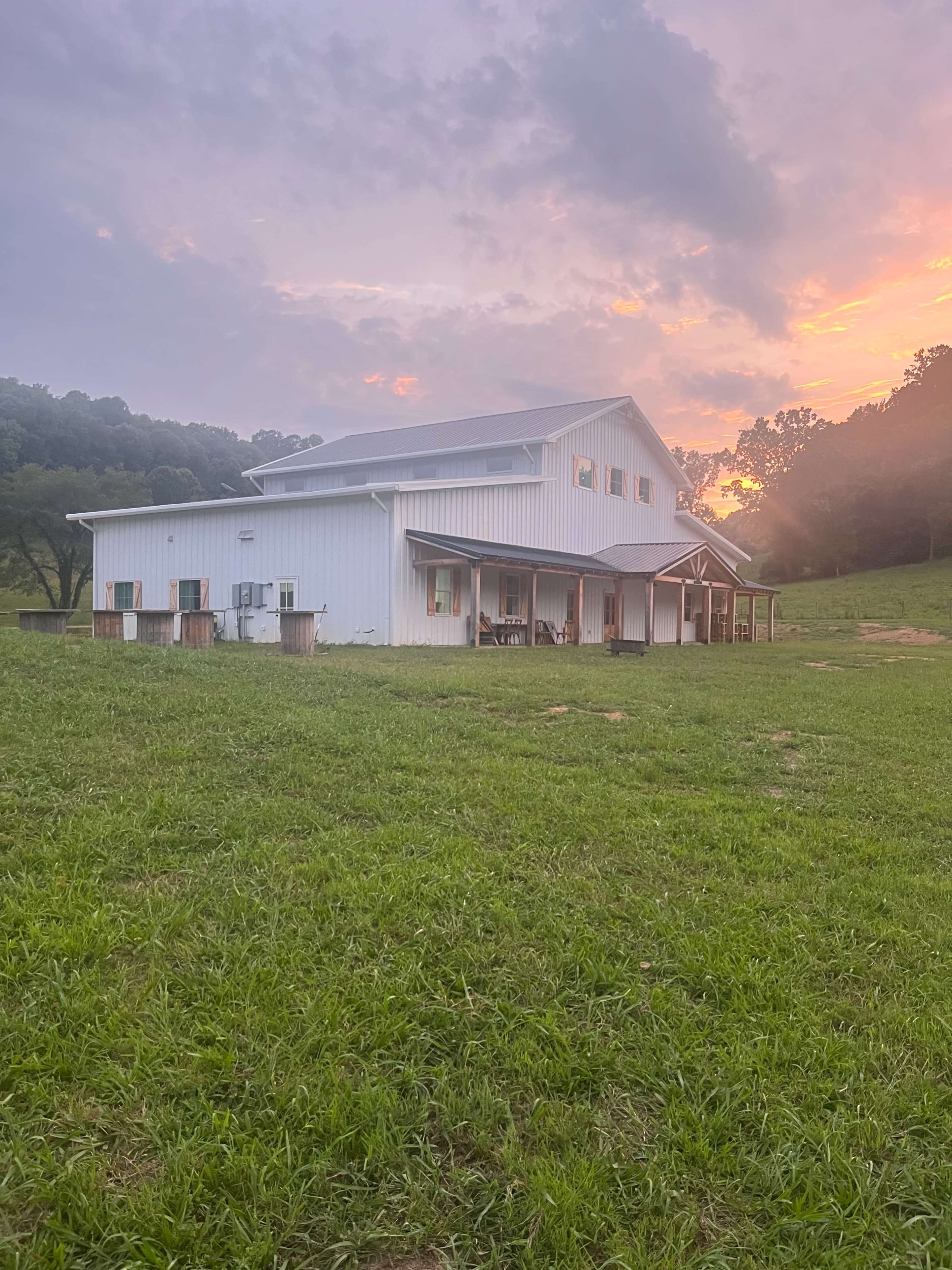 A large white barn is situated on a grassy field under a colorful sky at sunset.