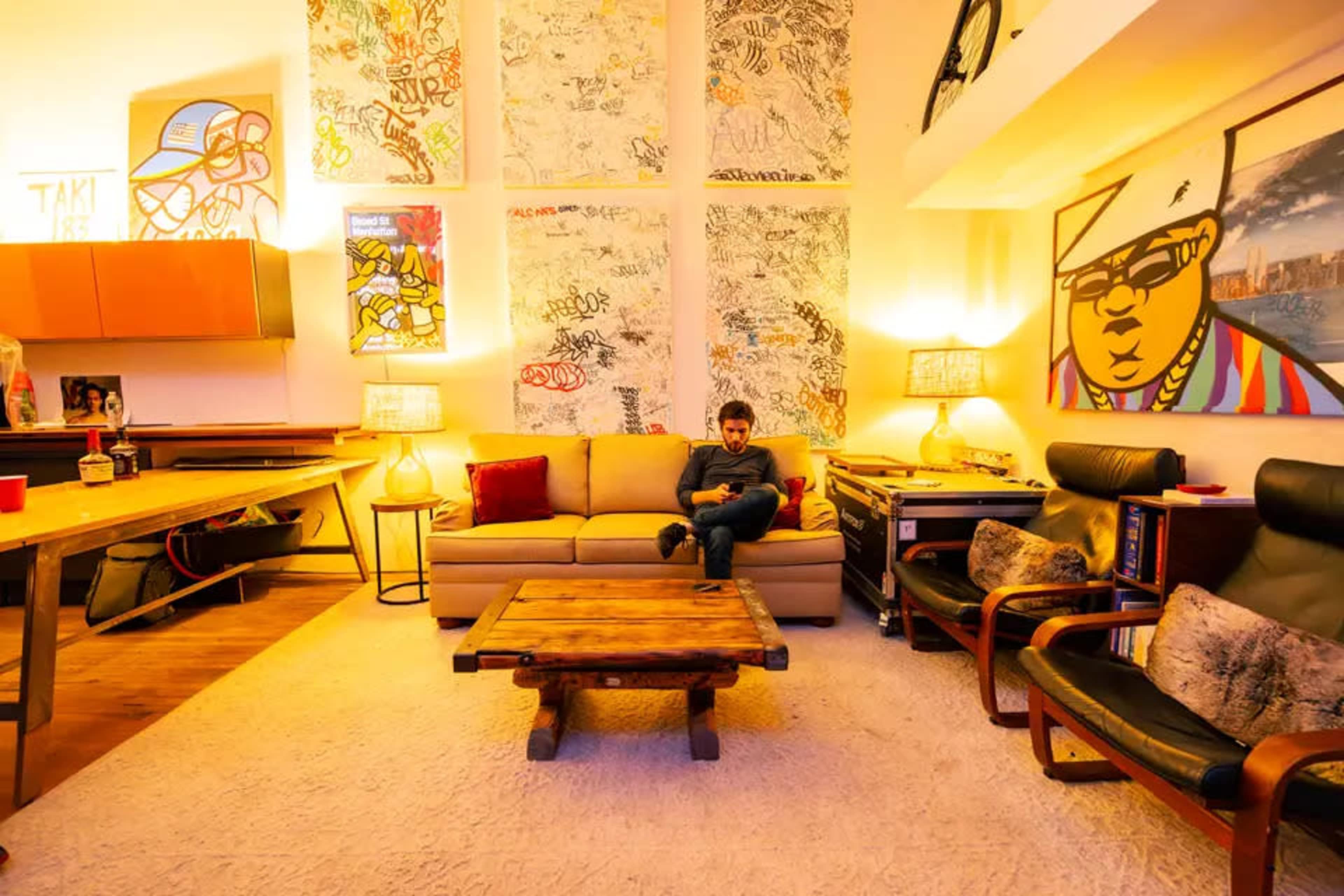 A person sits on a couch in a colorful room adorned with graffiti art on the walls, surrounded by wooden furniture and warm lighting.