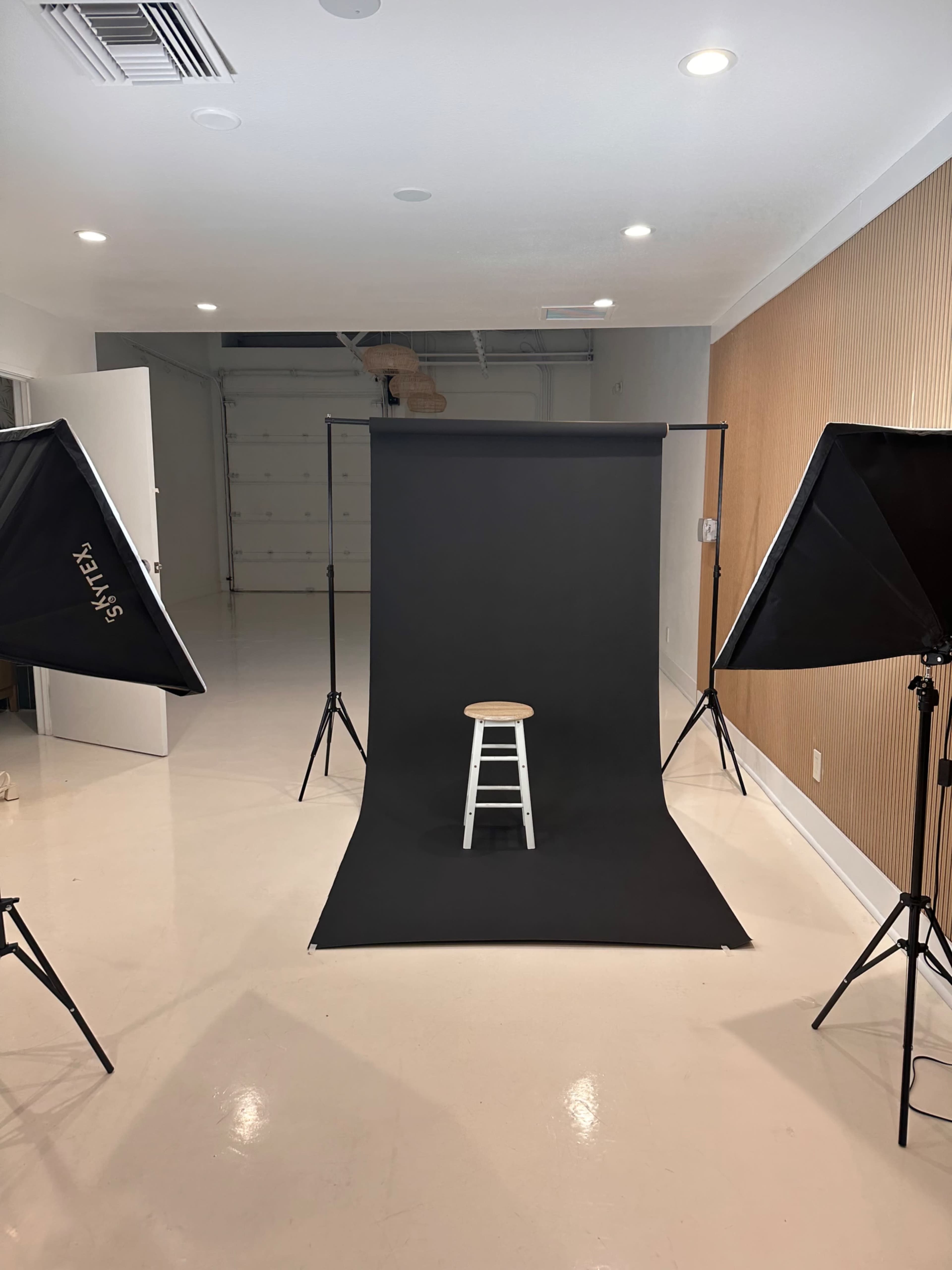 The image shows a photo studio setup featuring a black backdrop, a white stool in the center, and two lighting softboxes positioned on either side.