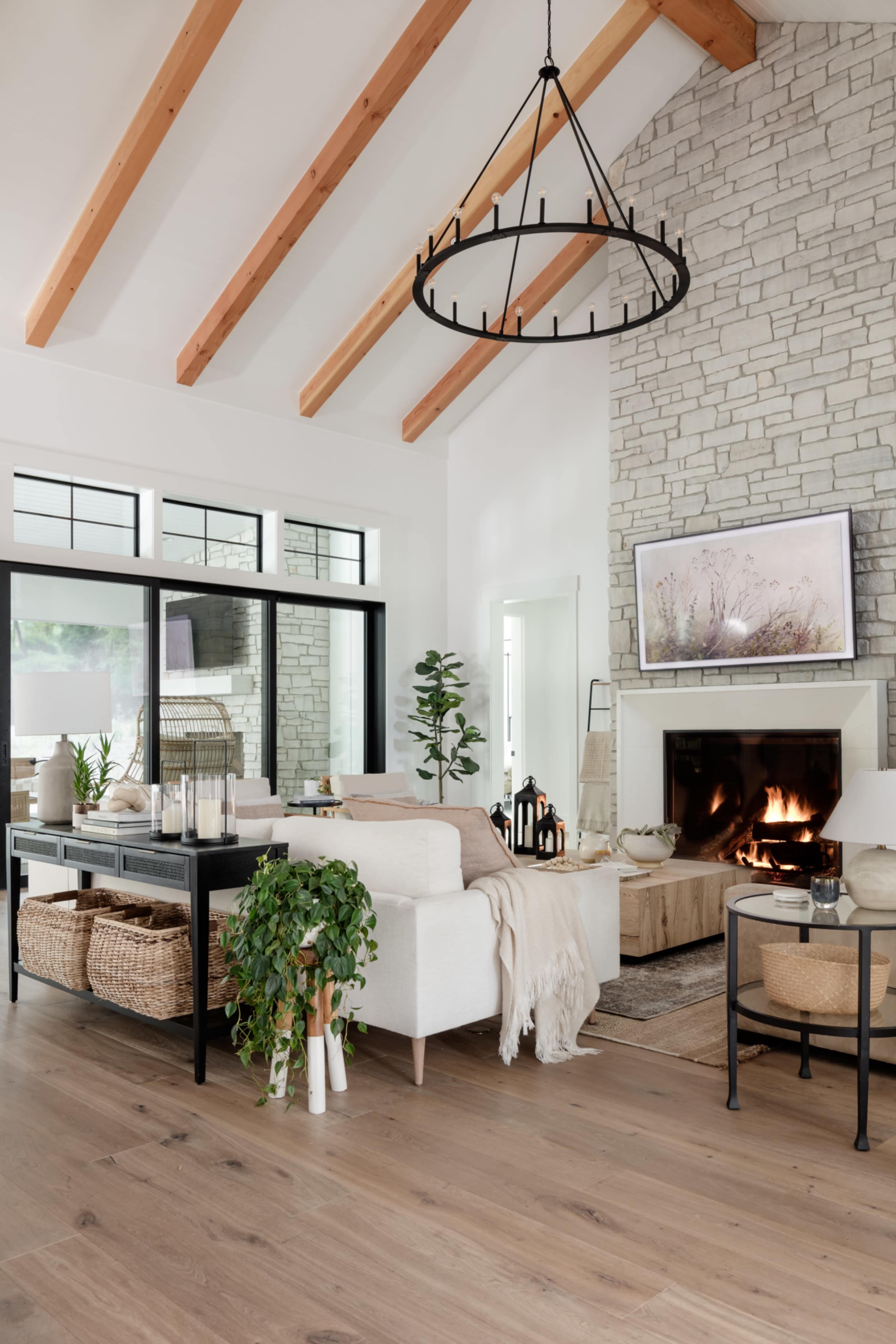 A modern living room features a stone fireplace, high wooden beams, and a white sofa adorned with a throw blanket, alongside decorative elements like plants and woven baskets.