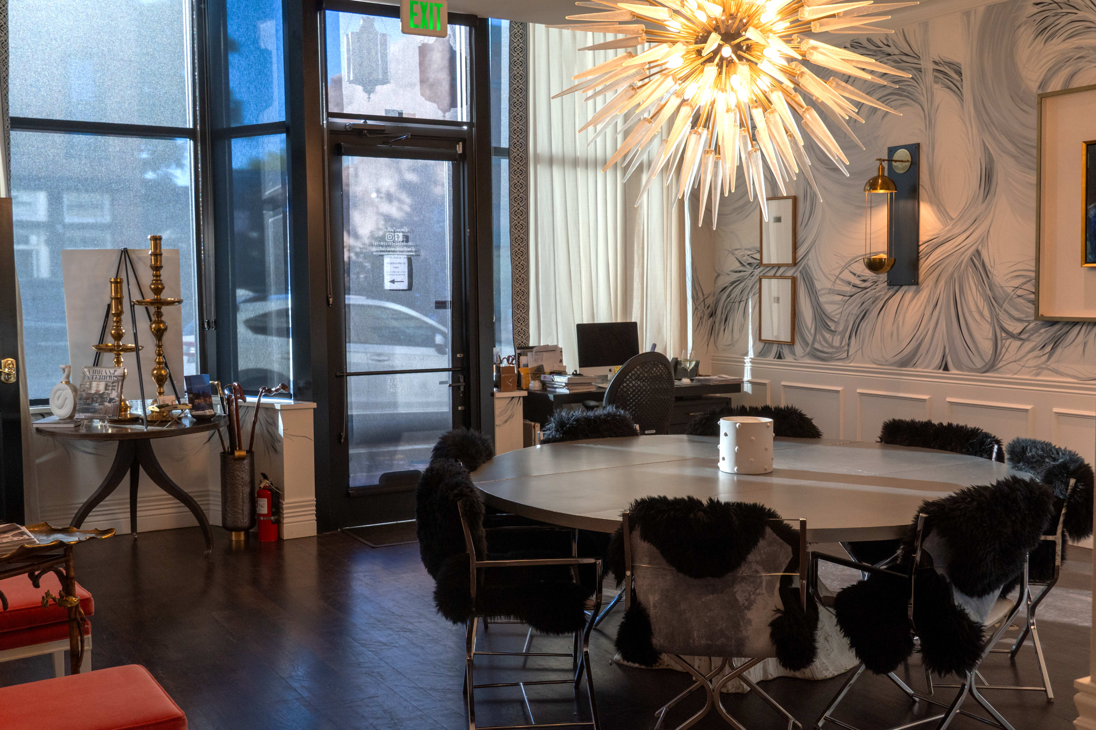 The image shows a modern office space with a round table surrounded by chairs, a large starburst chandelier overhead, and a decorative wall featuring abstract patterns.