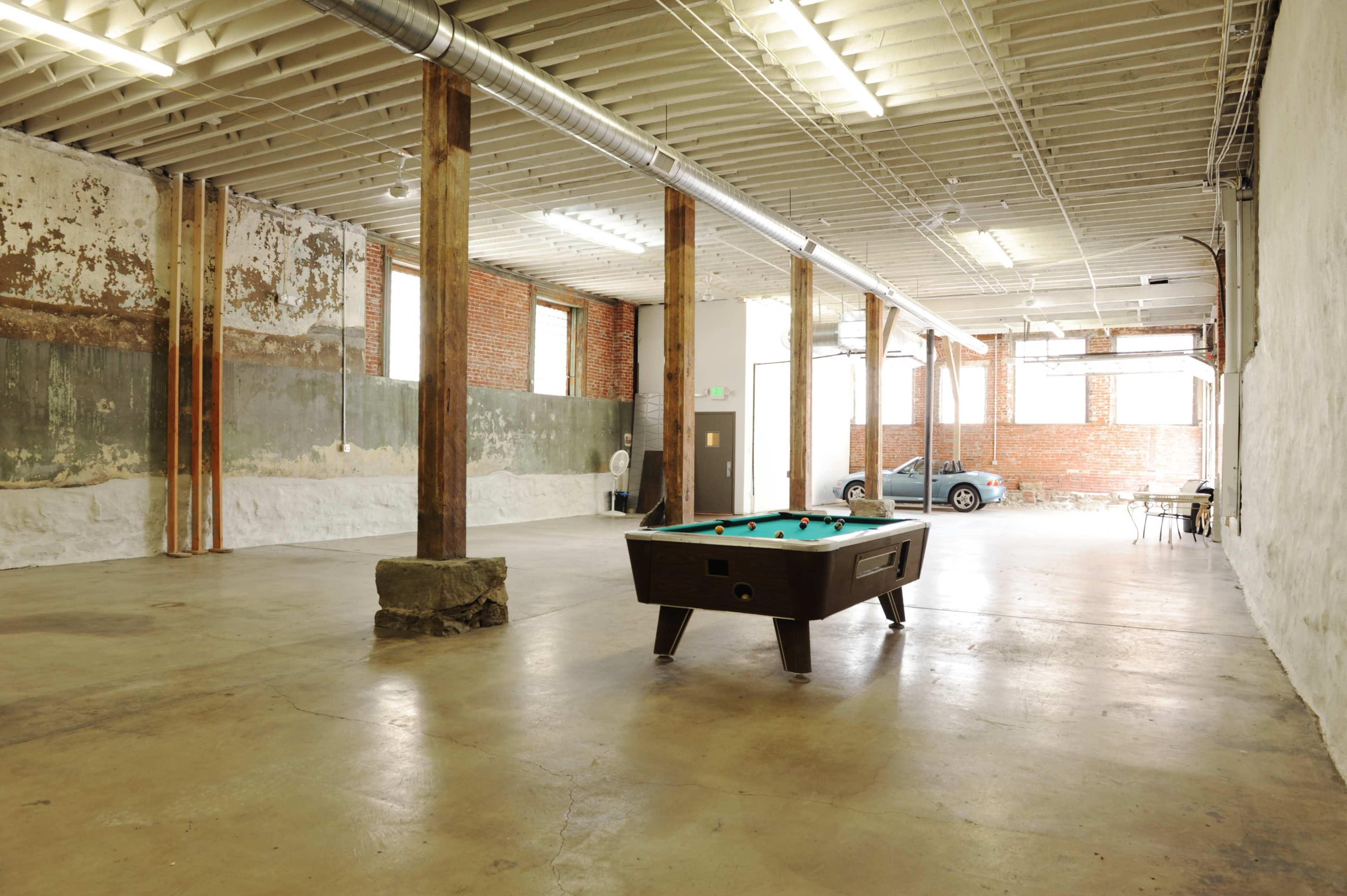 The image shows an empty industrial-style room with exposed brick walls, wooden columns, and a pool table in the center.