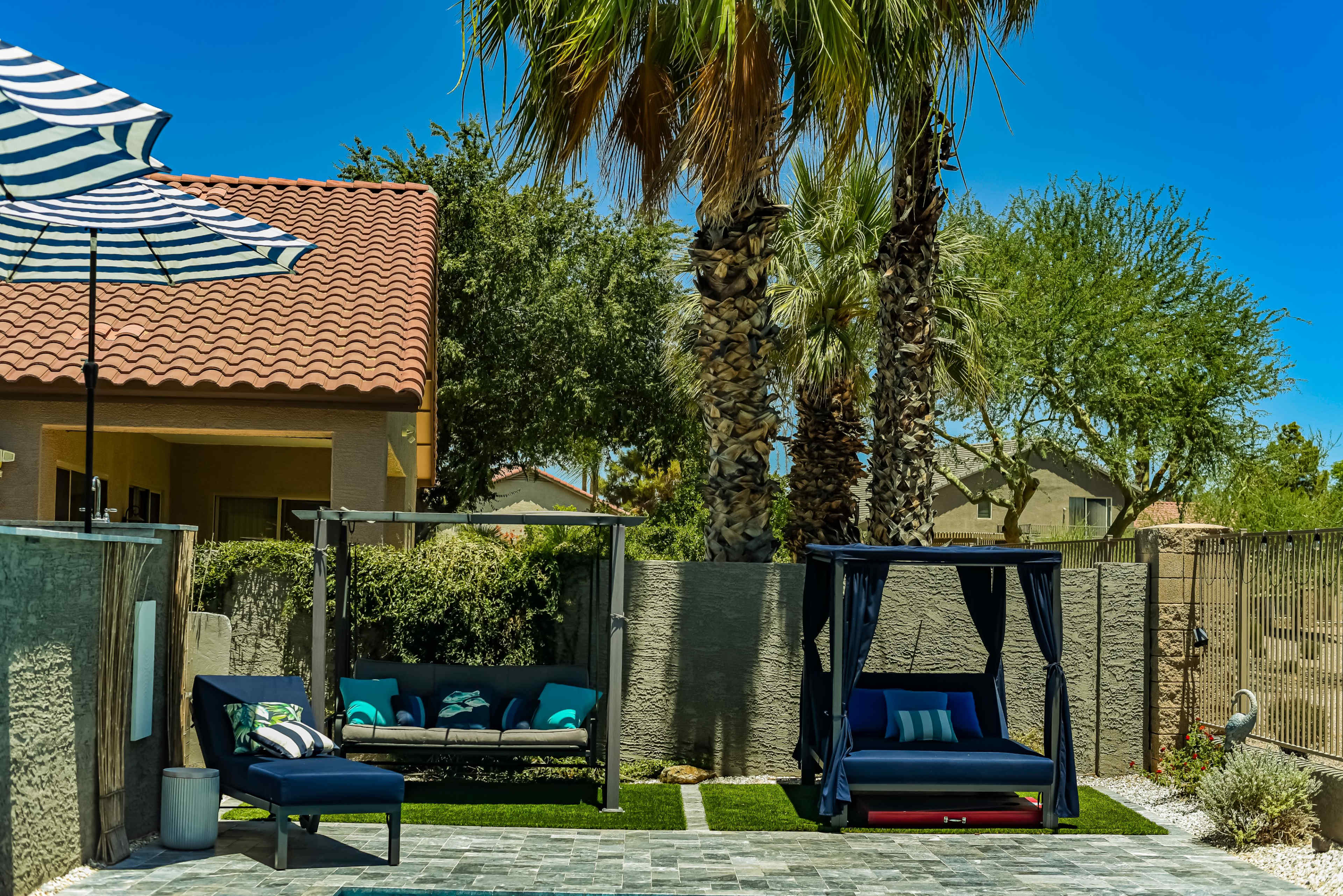 A patio area with outdoor furniture, including a blue sofa and a canopy swing, surrounded by palm trees and a shaded umbrella.