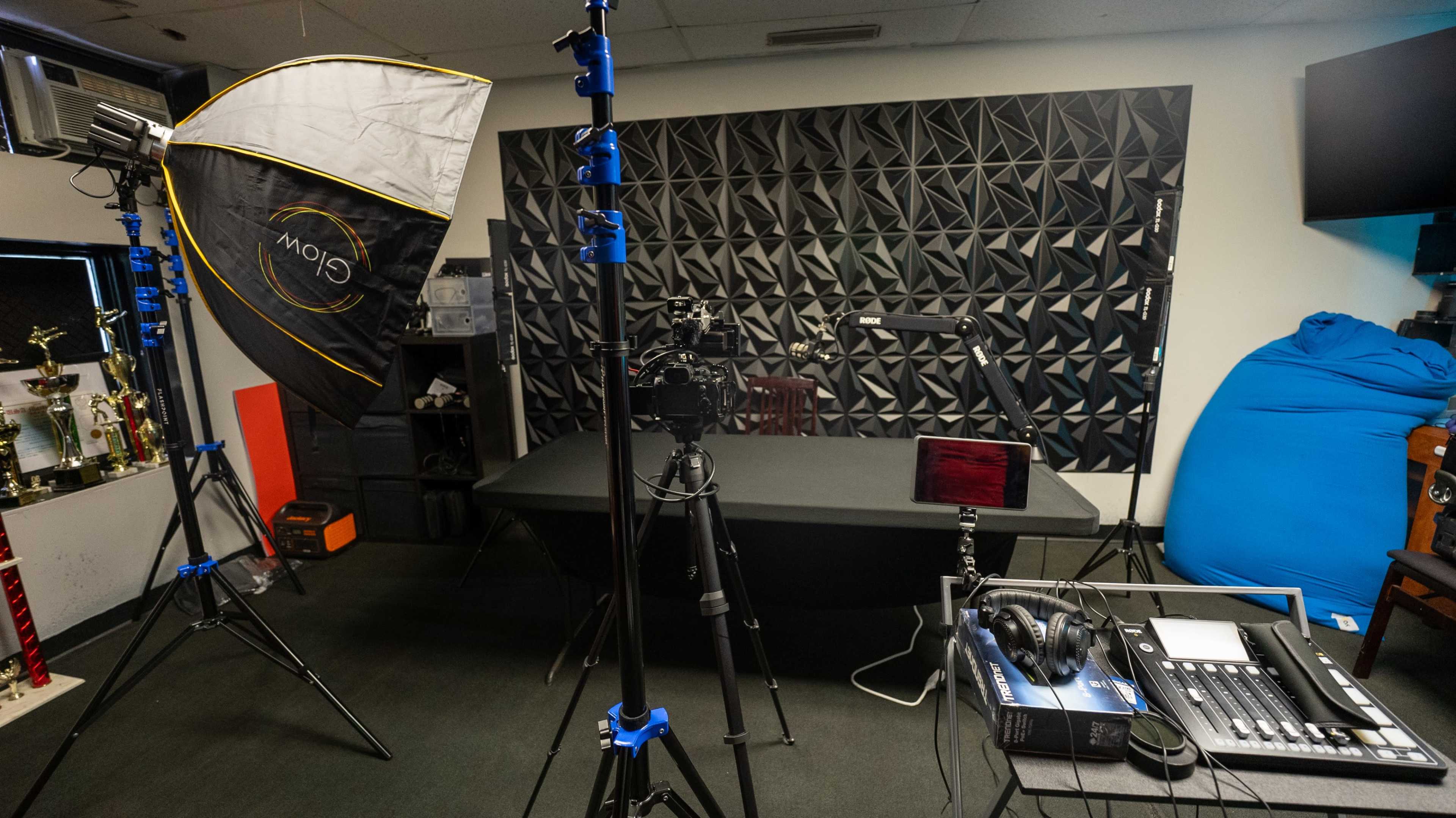 The image shows a recording studio equipped with lighting, cameras, and audio equipment, featuring a geometric soundproof wall and a large table.