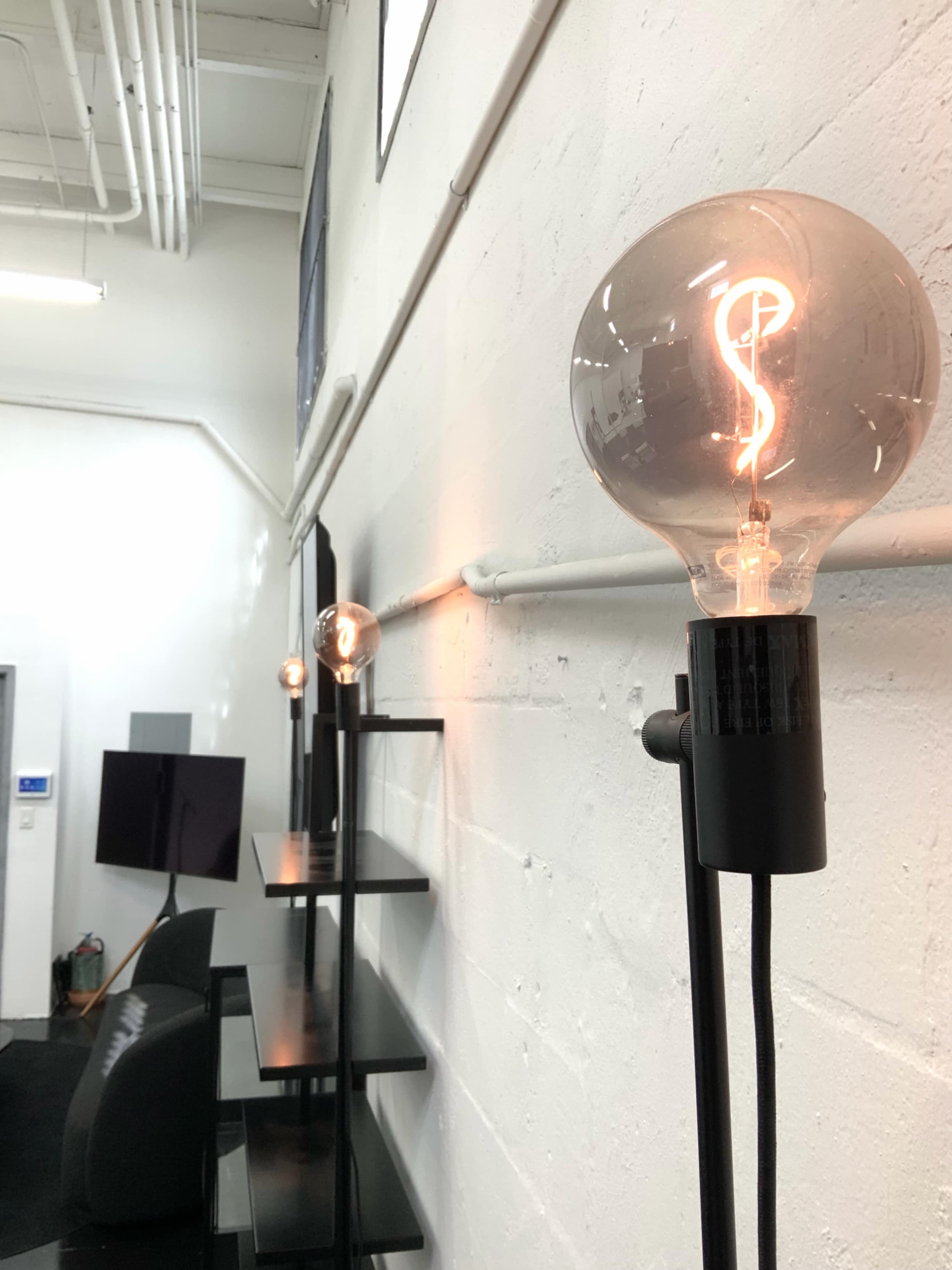 The image shows a row of illuminated, vintage-style light bulbs mounted on a white wall, with a modern shelving unit and a television in the background.