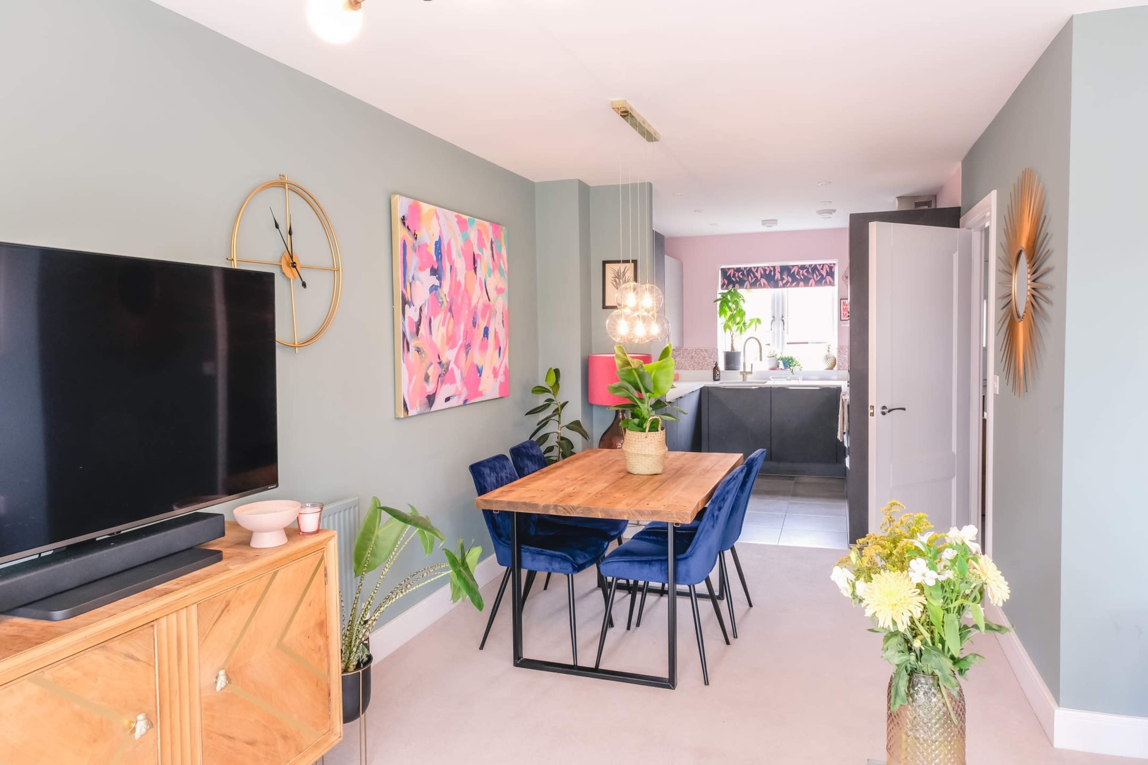 A modern dining area features a wooden table surrounded by blue chairs, with a television mounted on the wall and an open kitchen visible in the background.
