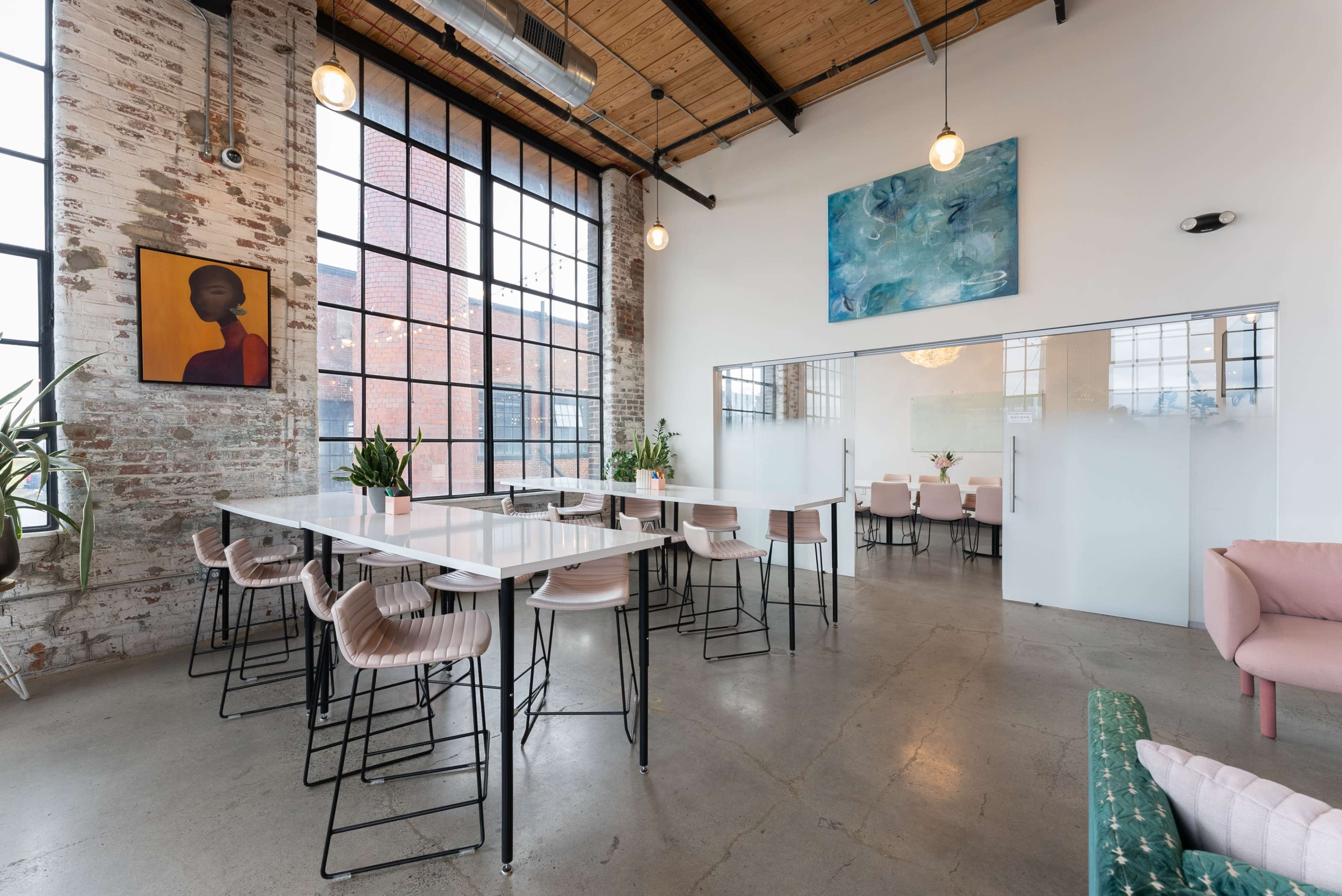 A spacious, modern interior with large windows, a long white table, and a mix of seating options, including pink chairs and a green couch.