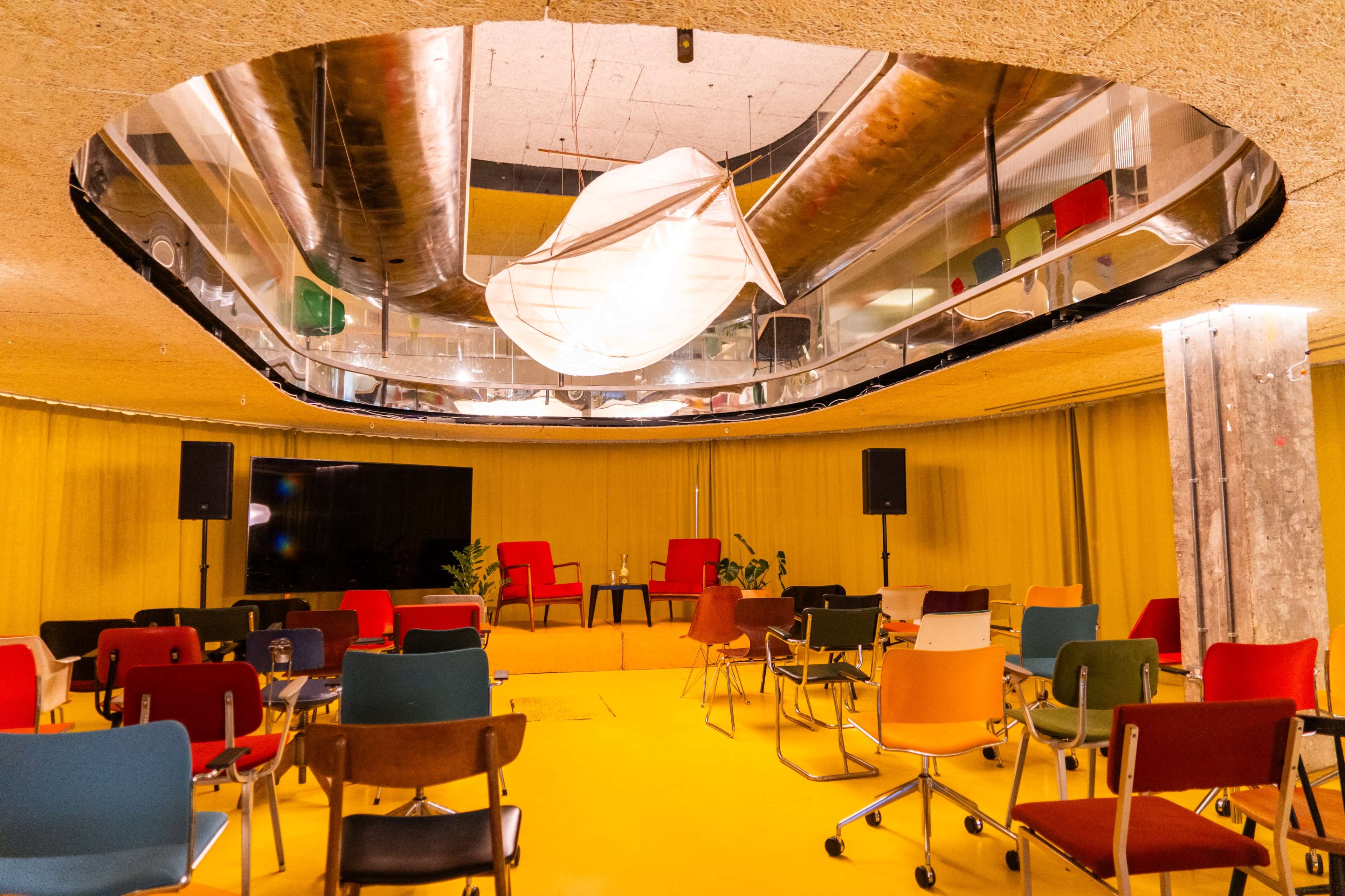 A modern event space features colorful chairs arranged in a circular pattern beneath a reflective ceiling and a large screen at the front.