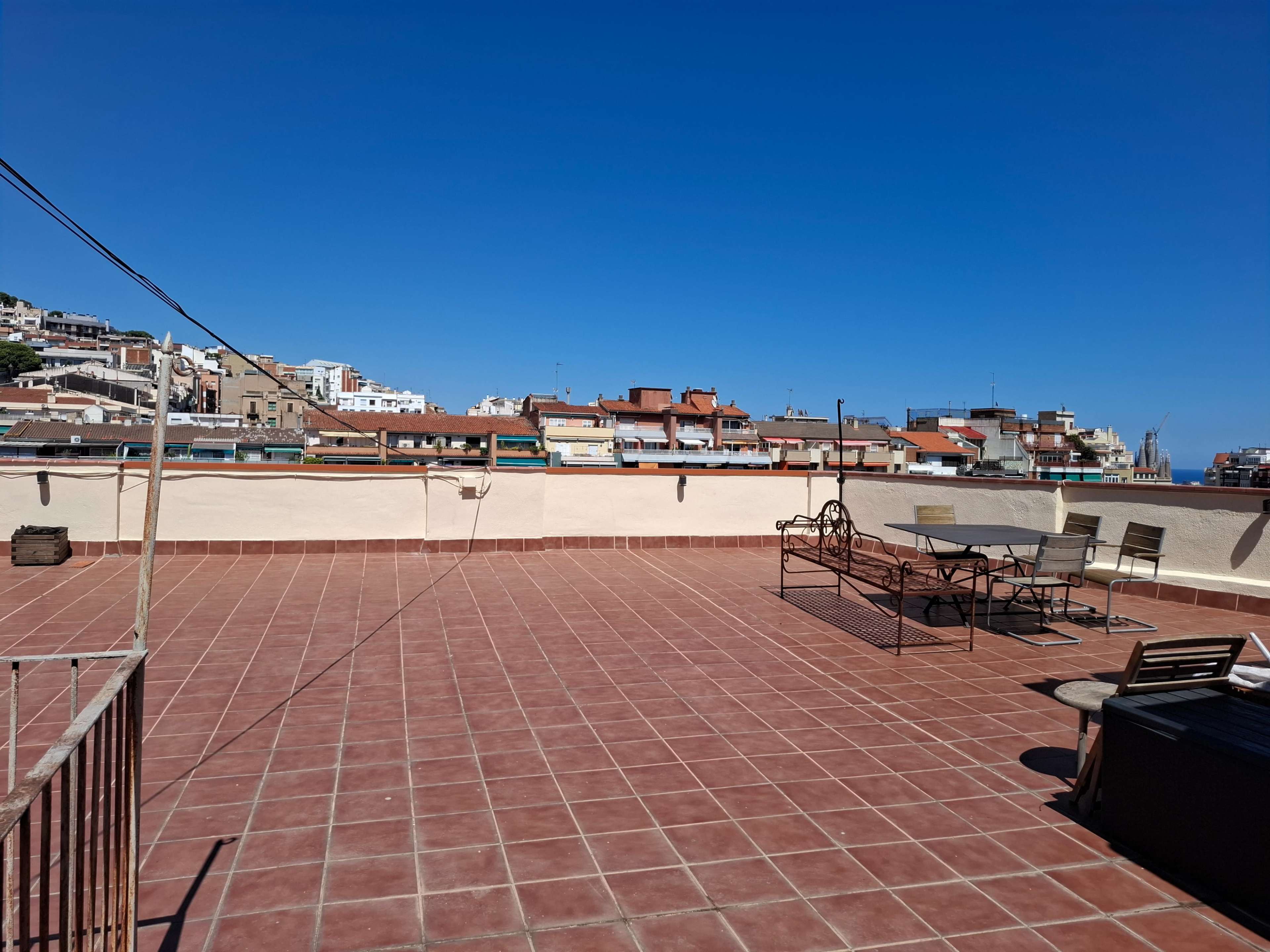The image shows a spacious rooftop terrace with a tiled floor and scattered furniture, overlooking a cityscape under a clear blue sky.